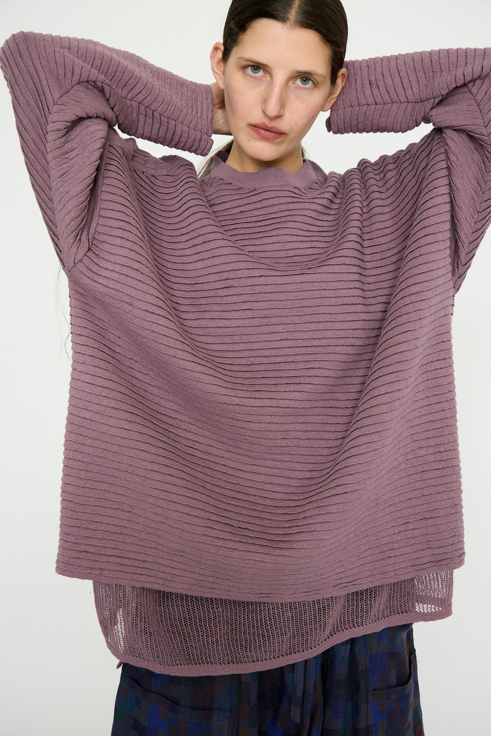 A person, wearing Jan-Jan Van Essche's Linen Cotton Yarn Knit #70 in Lavender, stands in patterned pants with their hands raised behind their head.