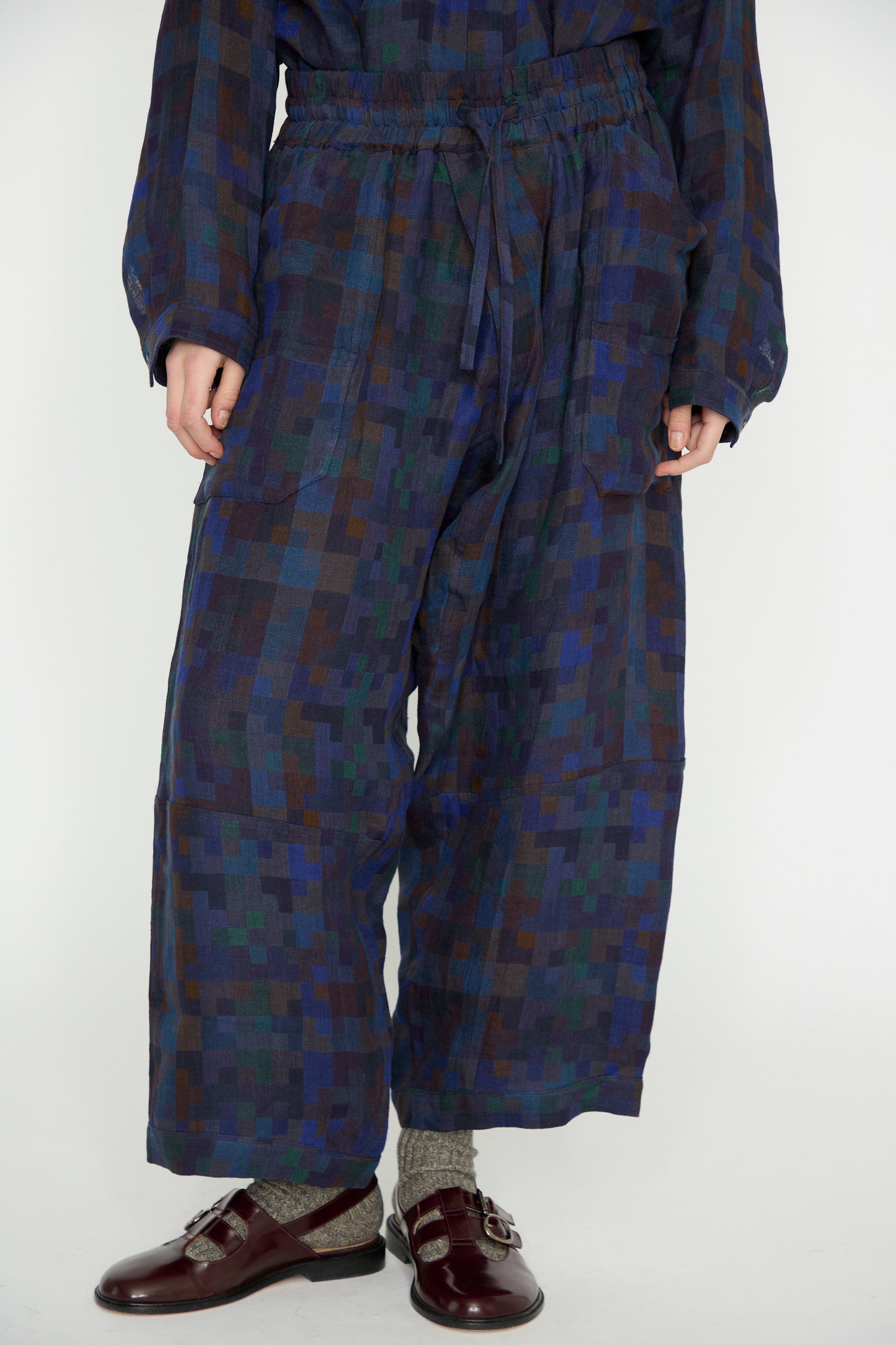 A person stands on a light background in Jan-Jan Van Essche's Linen Double Cloth Trouser #86 in Puzzle, featuring a blue and brown checkered design with loose-fit and elastic waist. The look is completed by brown shoes and gray socks, combining comfort with elegance seamlessly.