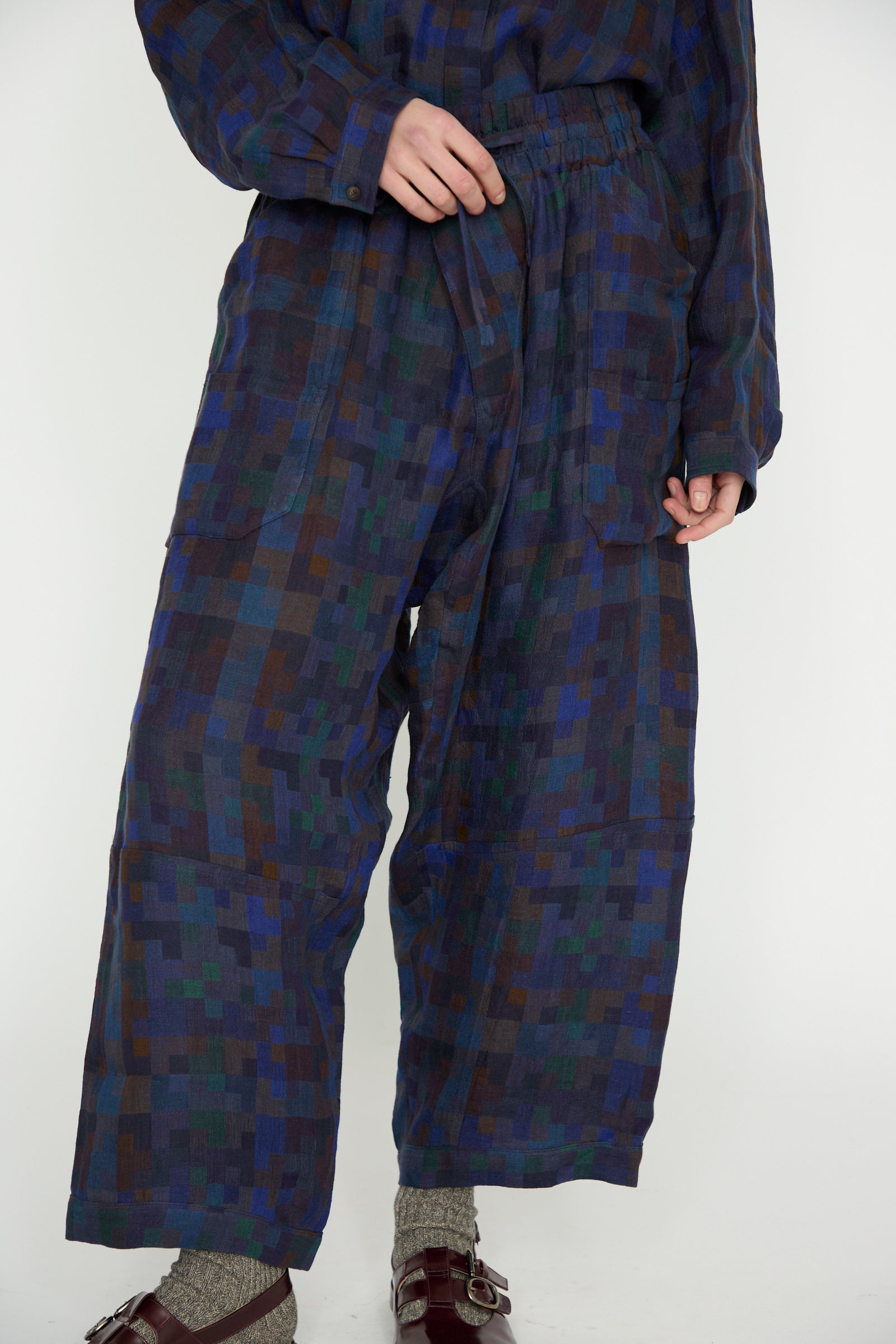 A person strikes a pose in a blue and green patterned outfit with Jan-Jan Van Essche's Linen Double Cloth Trouser #86 in Puzzle featuring loose-fit trousers with an elasticated waist, complemented by buckled sandals, against a plain background.