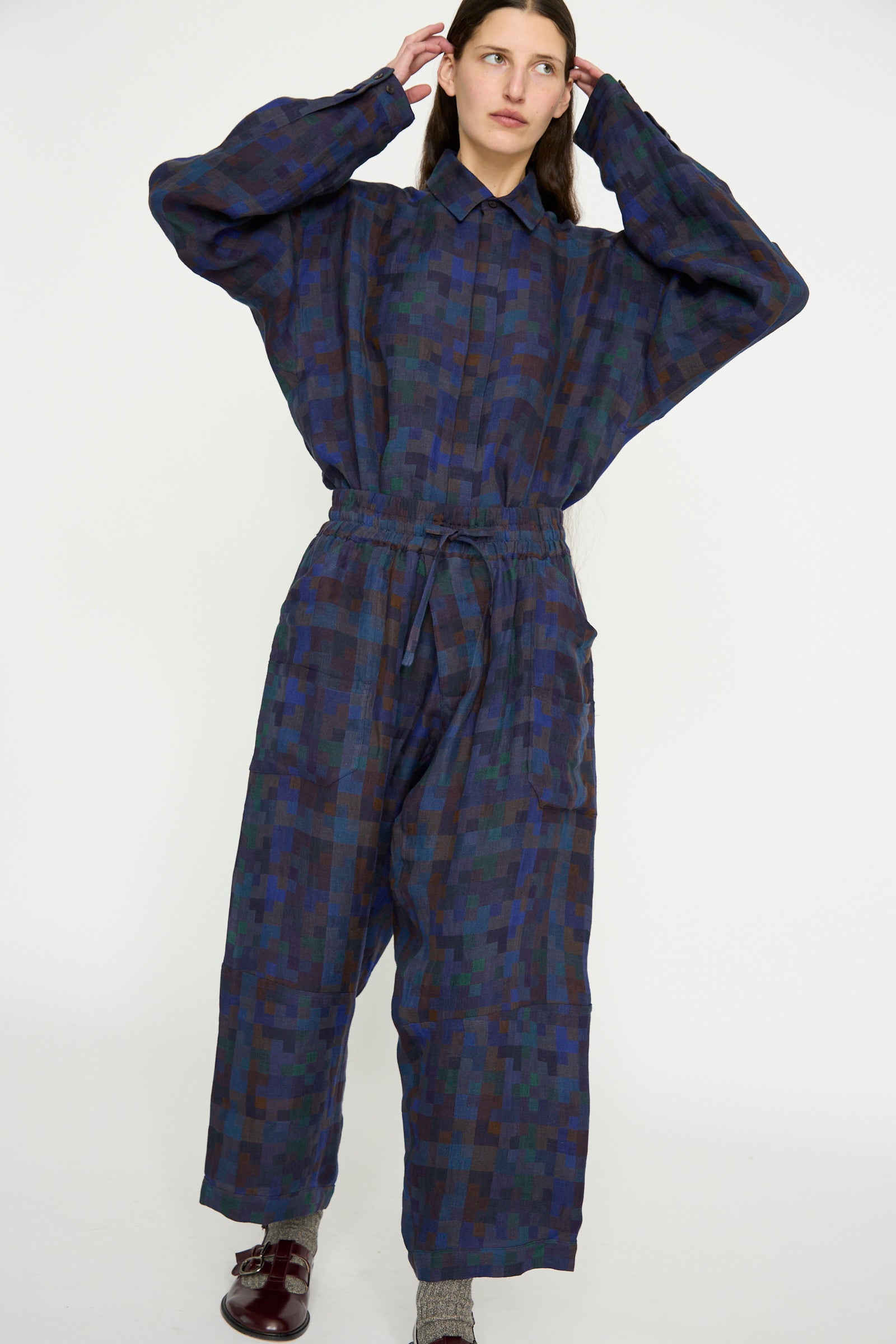 A person wearing the blue and green Jan-Jan Van Essche Linen Double Cloth Trouser #86 in Puzzle adjusts their hair with both hands. Featuring a collared jumpsuit style with loose-fit trousers, it offers comfort and style as they stand against a neutral background.