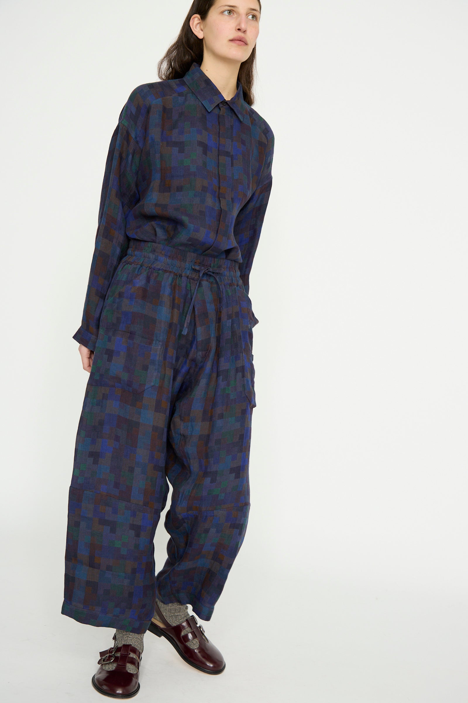 A person stands against a plain background wearing the Linen Double Cloth Trouser #86 in Puzzle by Jan-Jan Van Essche, featuring a matching blue and green checkered design. These wide-leg pants offer a loose fit with an elasticated waist, blending style and comfort effortlessly.