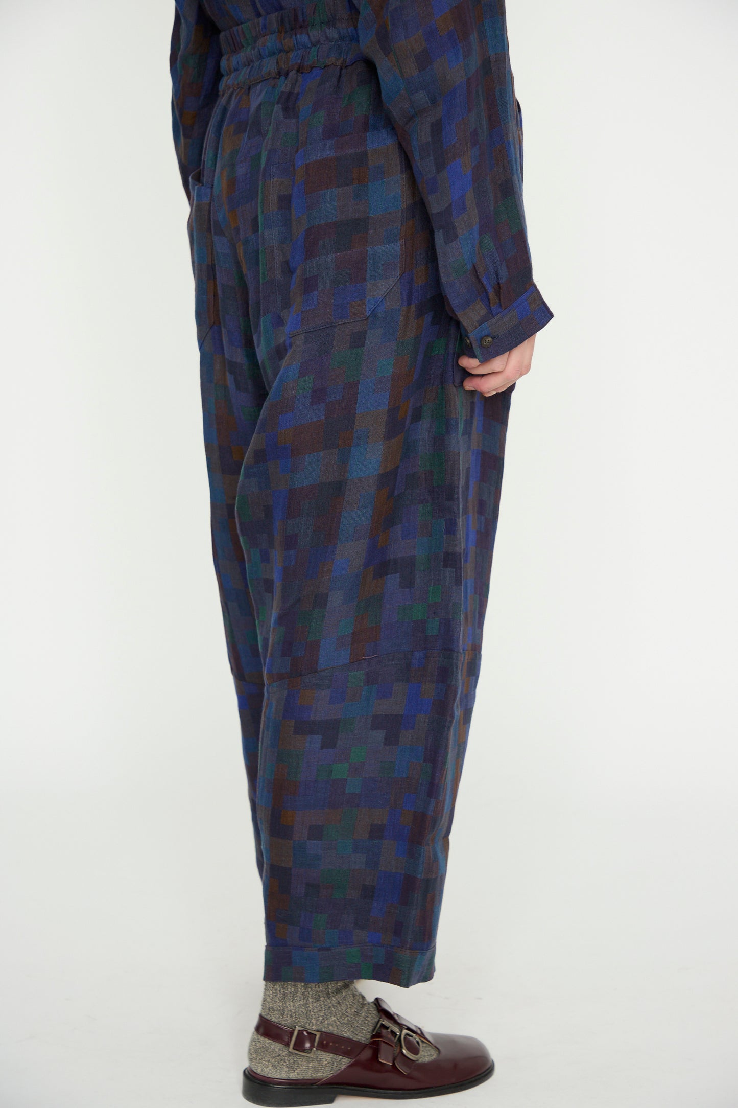 A person in Jan-Jan Van Essche's Linen Double Cloth Trouser #86 in Puzzle, featuring a checkered blue and green pattern and elasticated waist, stands sideways wearing maroon shoes and gray socks.