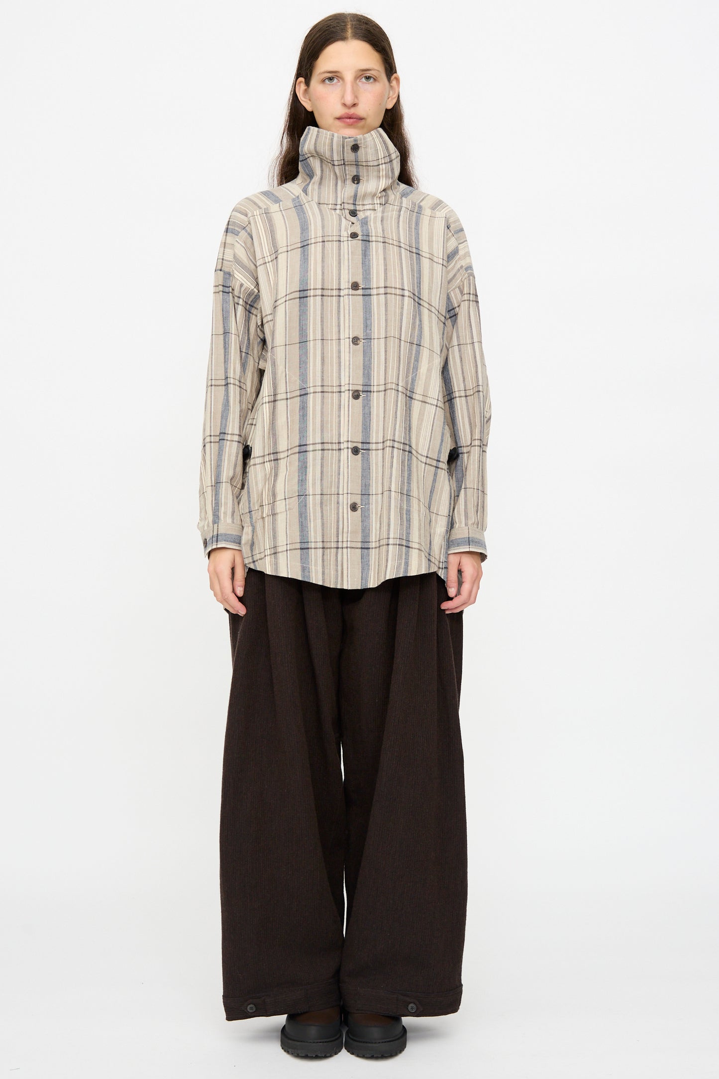 A person stands against a plain background wearing the Madras Plaid Shirt #110 in Plaster by Jan-Jan Van Essche, styled with wide-leg dark brown pants.