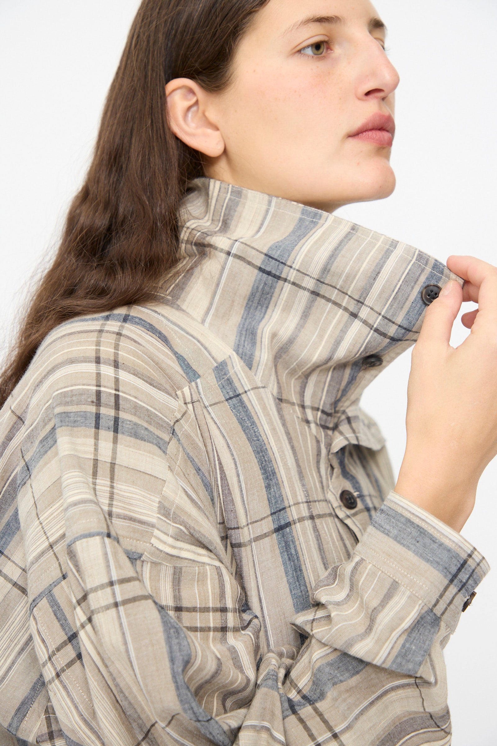 A person with long brown hair wears the Jan-Jan Van Essche Madras Plaid Shirt #110 in Plaster, featuring a beige and blue cotton blend plaid with the collar turned up, looking to the side against a plain background.