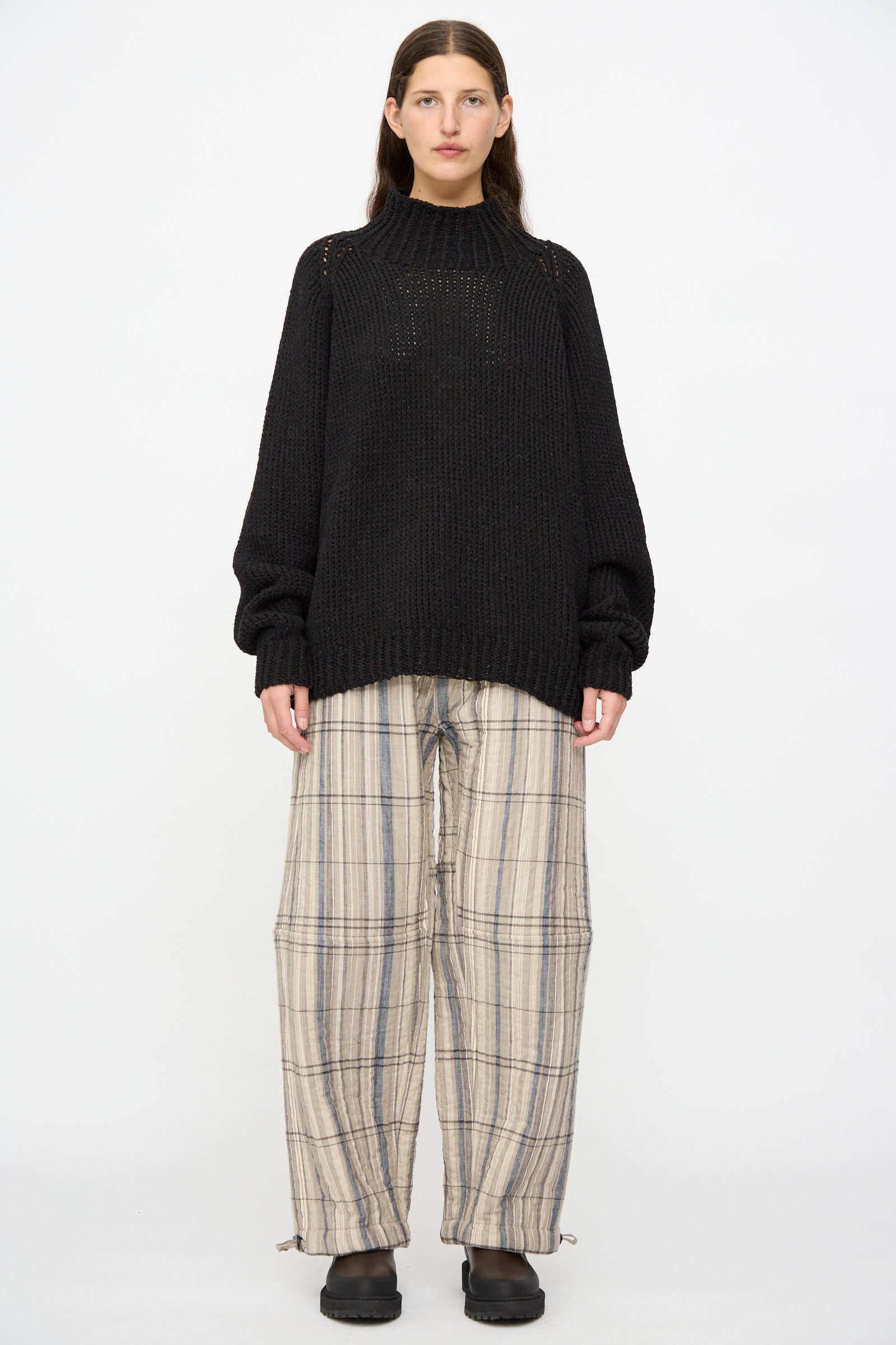 A person stands against a plain background wearing a black knit sweater, Jan-Jan Van Essche Madras Plaid Trouser #89 in Plaster, and black shoes.