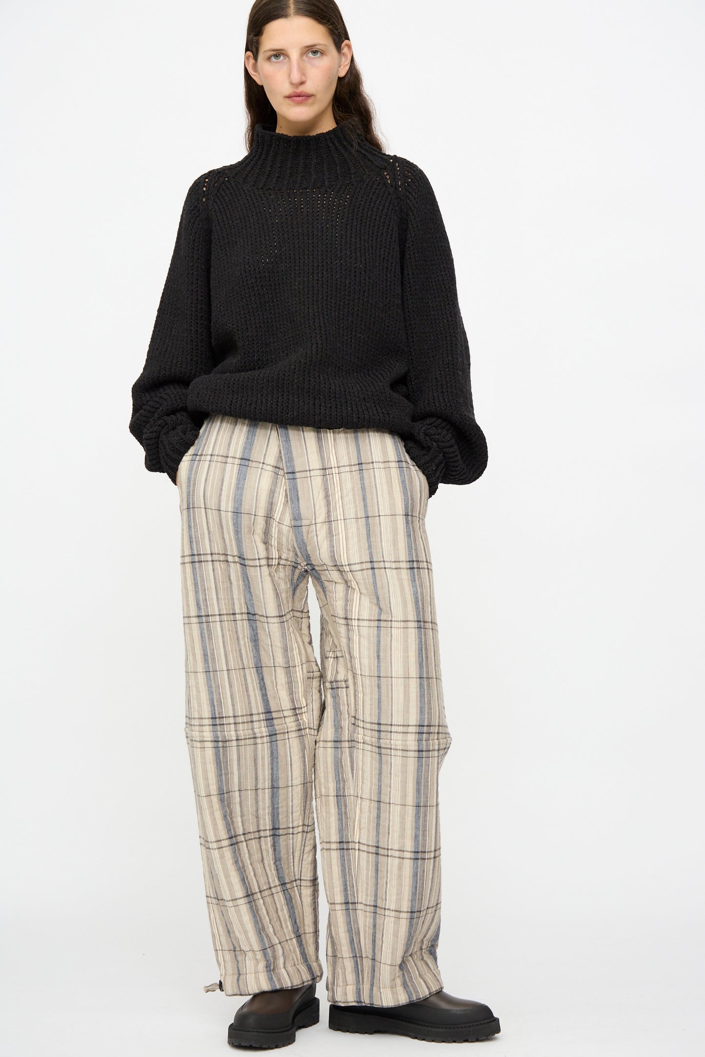 A person wears Jan-Jan Van Essche's Madras Plaid Trouser #89 in Plaster, a black knit sweater, and black shoes, standing against a plain white background.