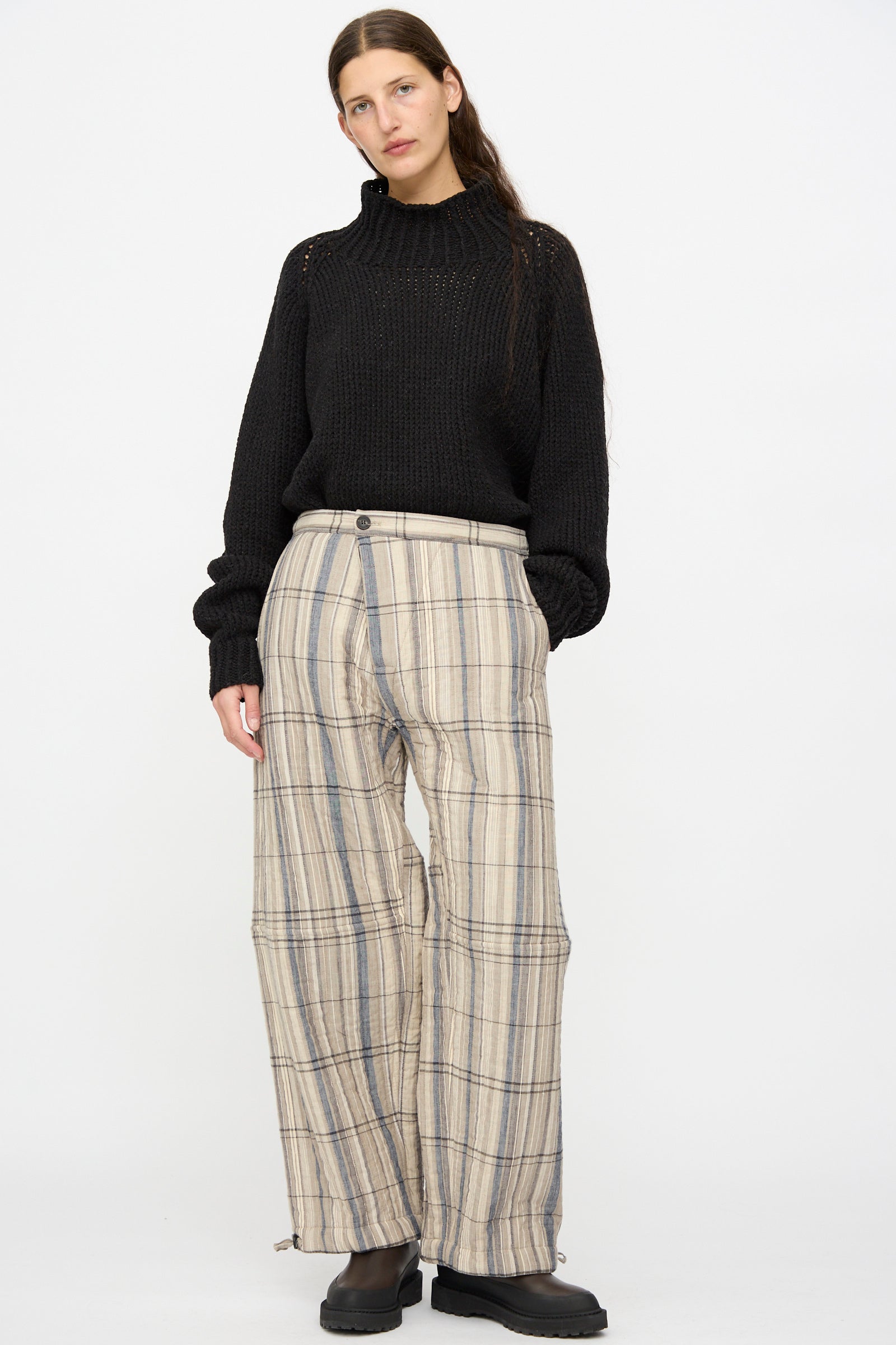 Person stands against a plain background wearing a black knit sweater, Jan-Jan Van Essche Madras Plaid Trouser #89 in Plaster, and black shoes.