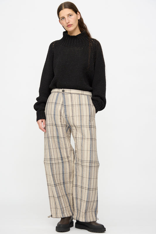 Person stands against a plain background wearing a black knit sweater, Jan-Jan Van Essche Madras Plaid Trouser #89 in Plaster, and black shoes.