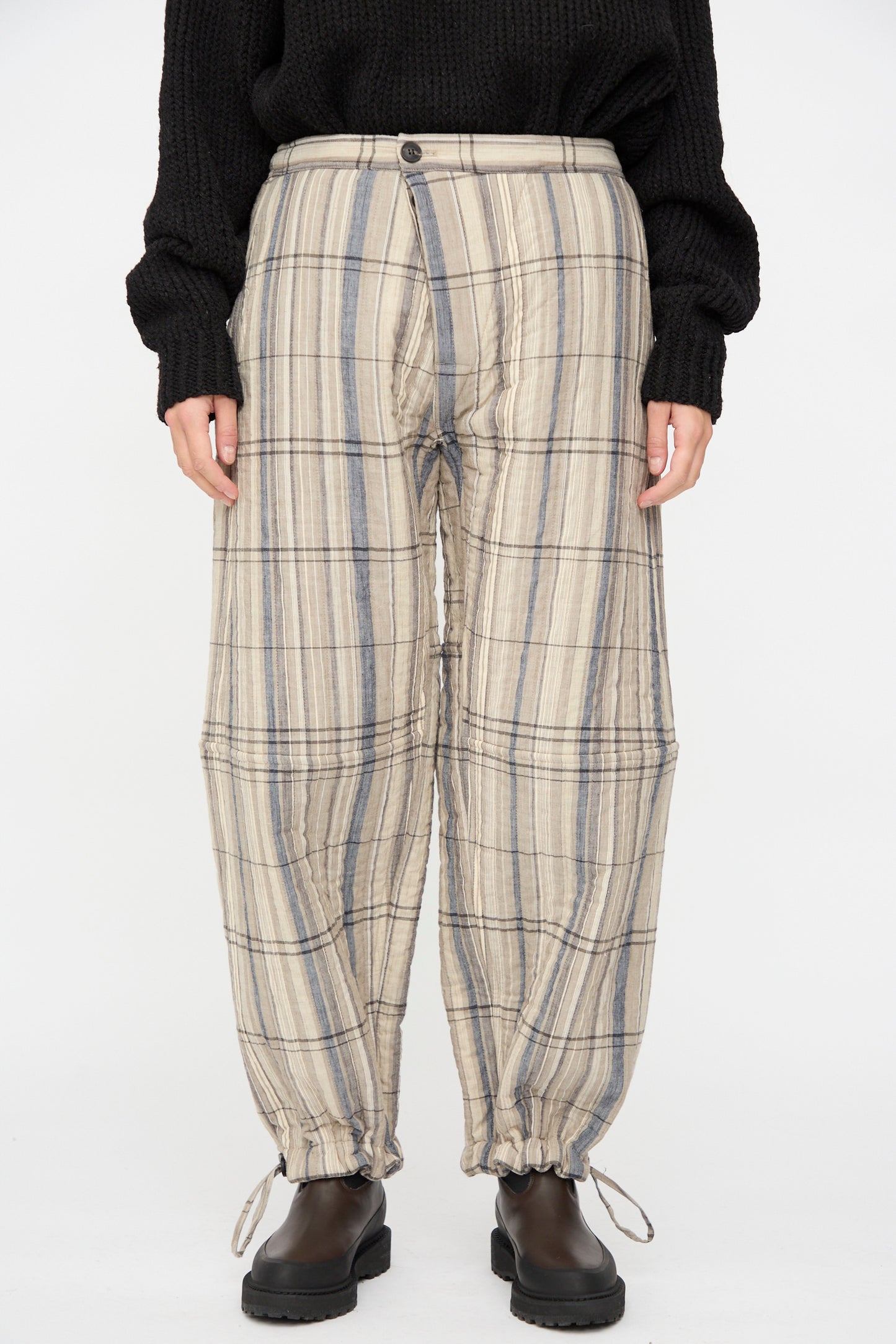A person wears the Jan-Jan Van Essche Madras Plaid Trouser #89 in Plaster—relaxed fit, beige and gray plaid cotton blend with drawstring hems—styled with a black sweater and boots, standing against a plain white background.