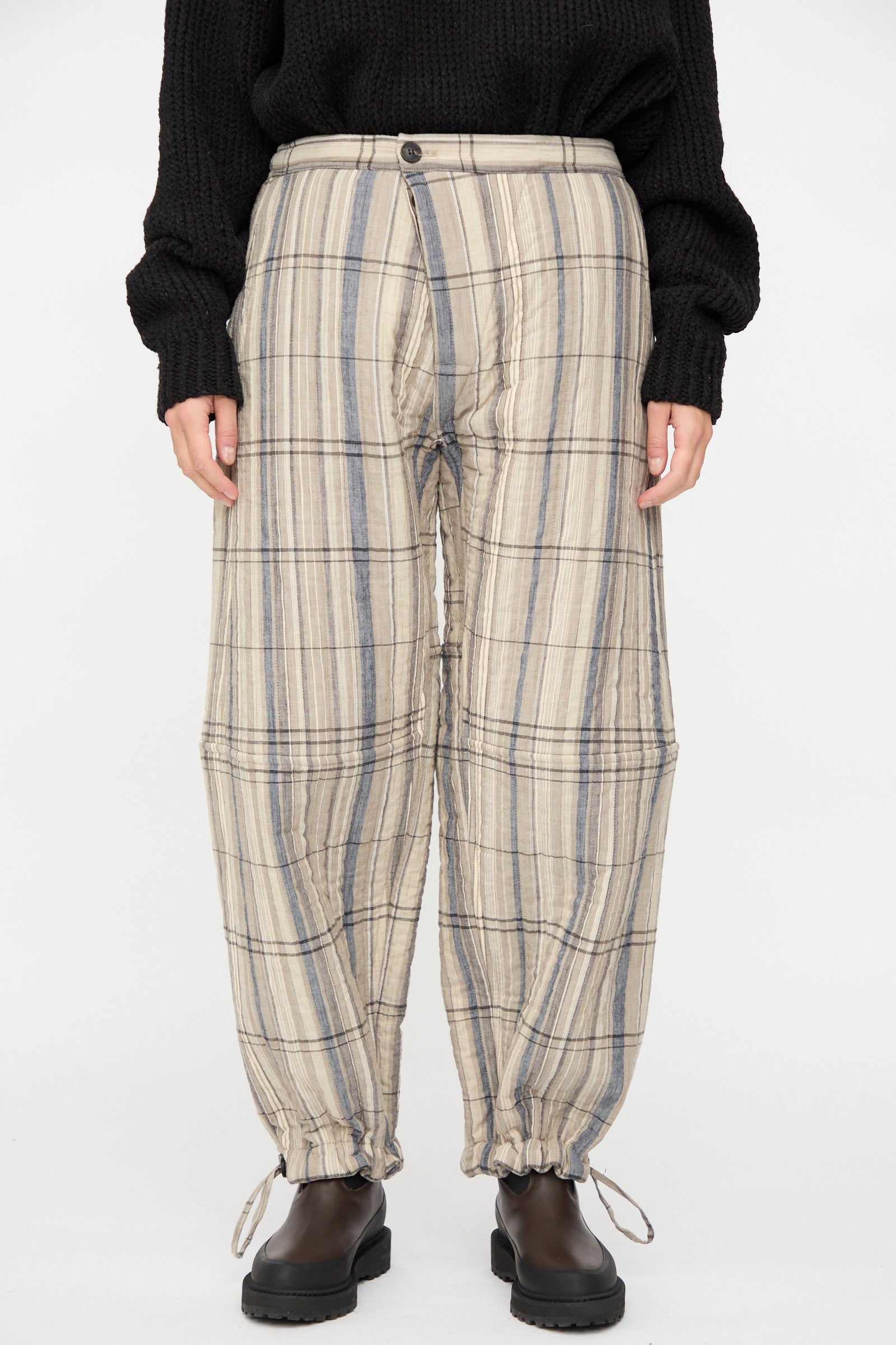 A person wears the Jan-Jan Van Essche Madras Plaid Trouser #89 in Plaster—relaxed fit, beige and gray plaid cotton blend with drawstring hems—styled with a black sweater and boots, standing against a plain white background.