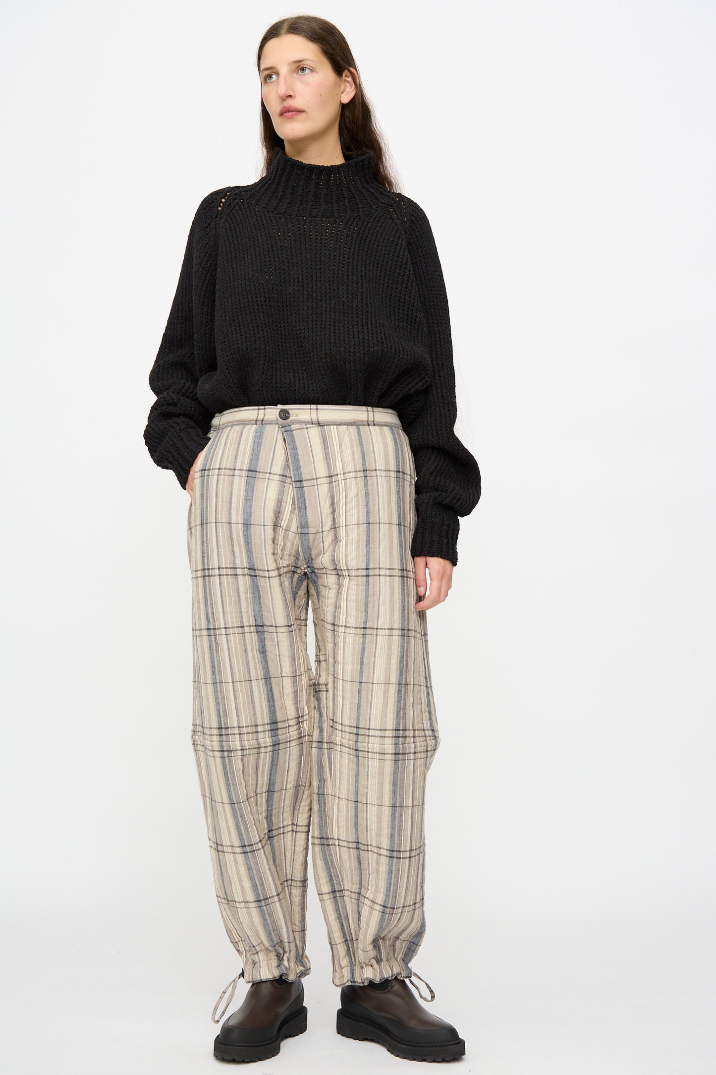 A woman stands against a plain background wearing a black knit sweater, black shoes, and Jan-Jan Van Essche’s Madras Plaid Trouser #89 in Plaster.
