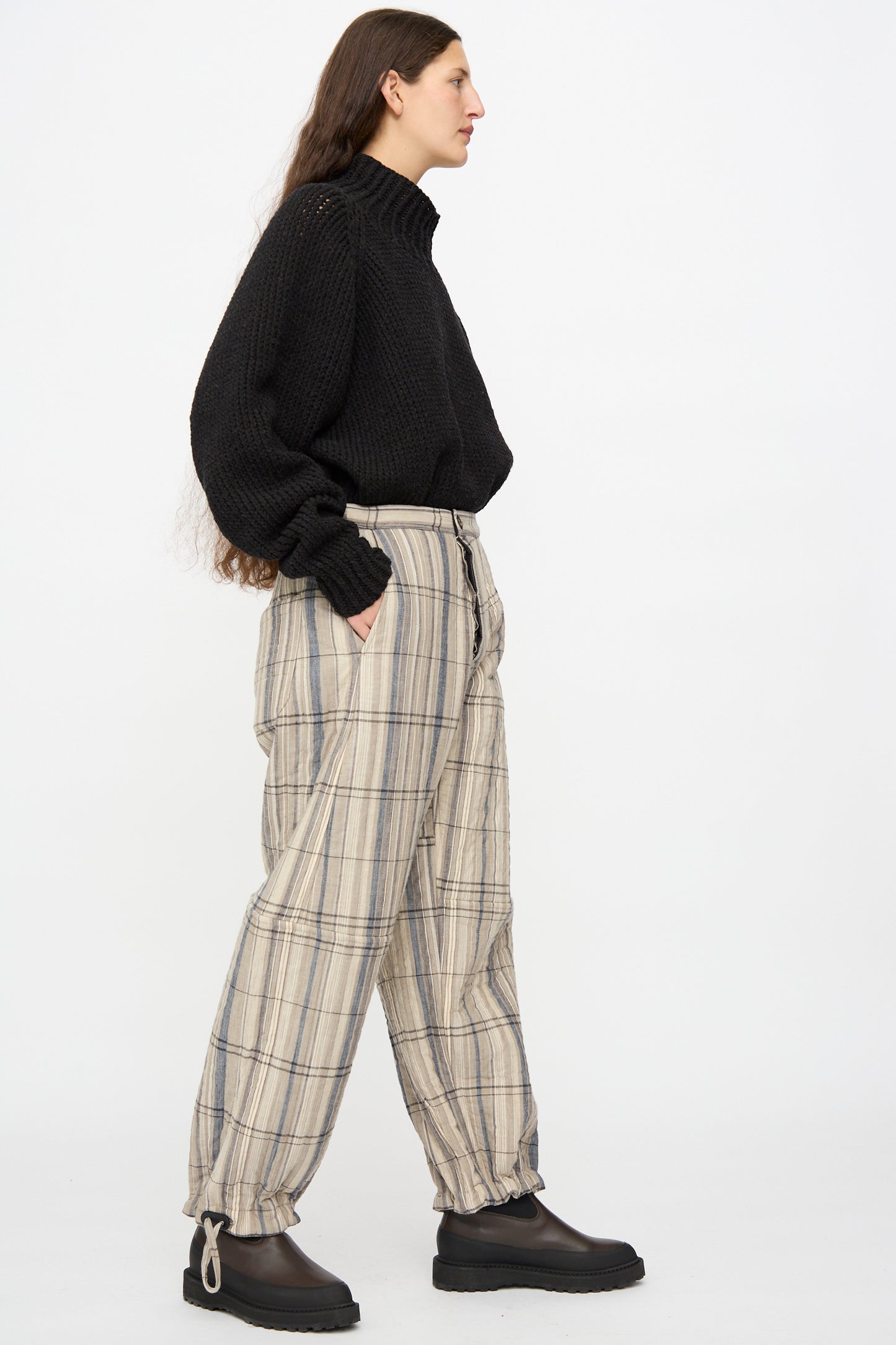 A person stands sideways against a white background wearing a black turtleneck, Jan-Jan Van Essche Madras Plaid Trouser #89 in Plaster, and black platform shoes.