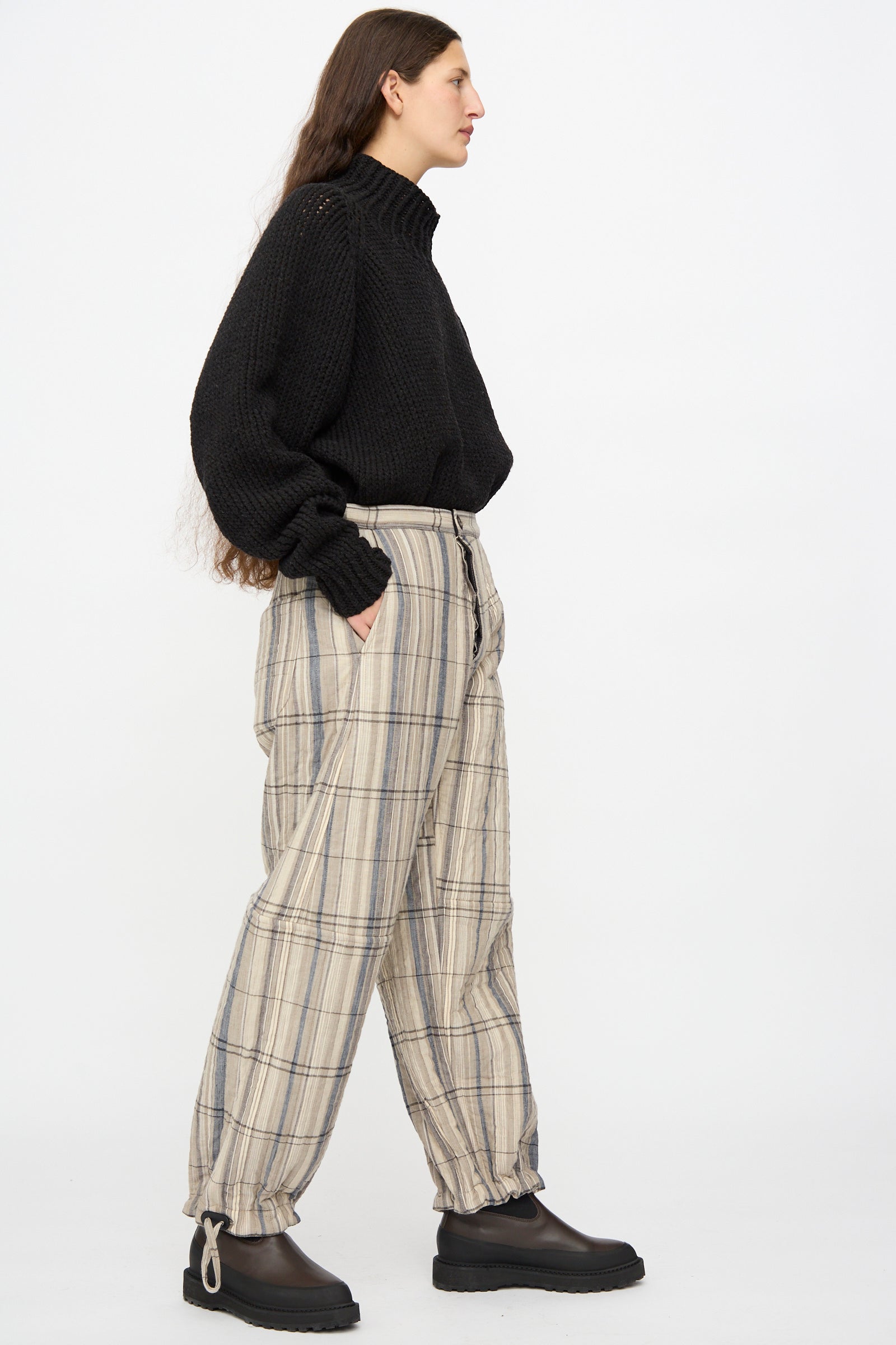 A person stands sideways against a white background wearing a black turtleneck, Jan-Jan Van Essche Madras Plaid Trouser #89 in Plaster, and black platform shoes.