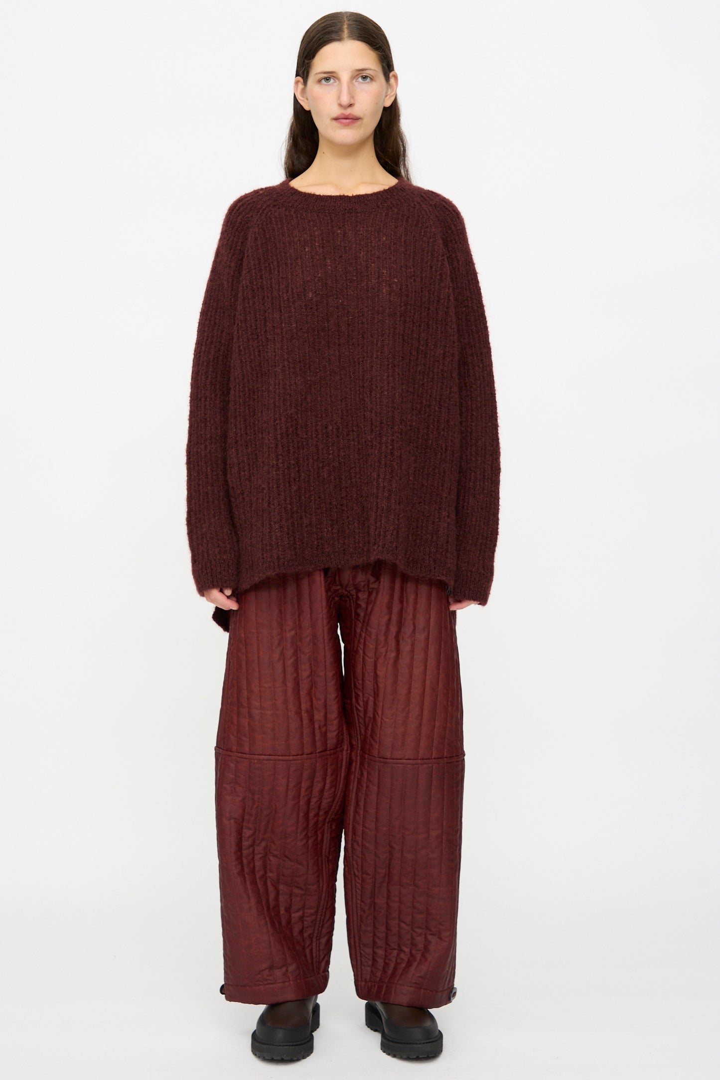A person stands against a white background wearing the Jan-Jan Van Essche Mohair Knit #71 Sweater in Oxblood, loose quilted maroon pants, and black shoes.