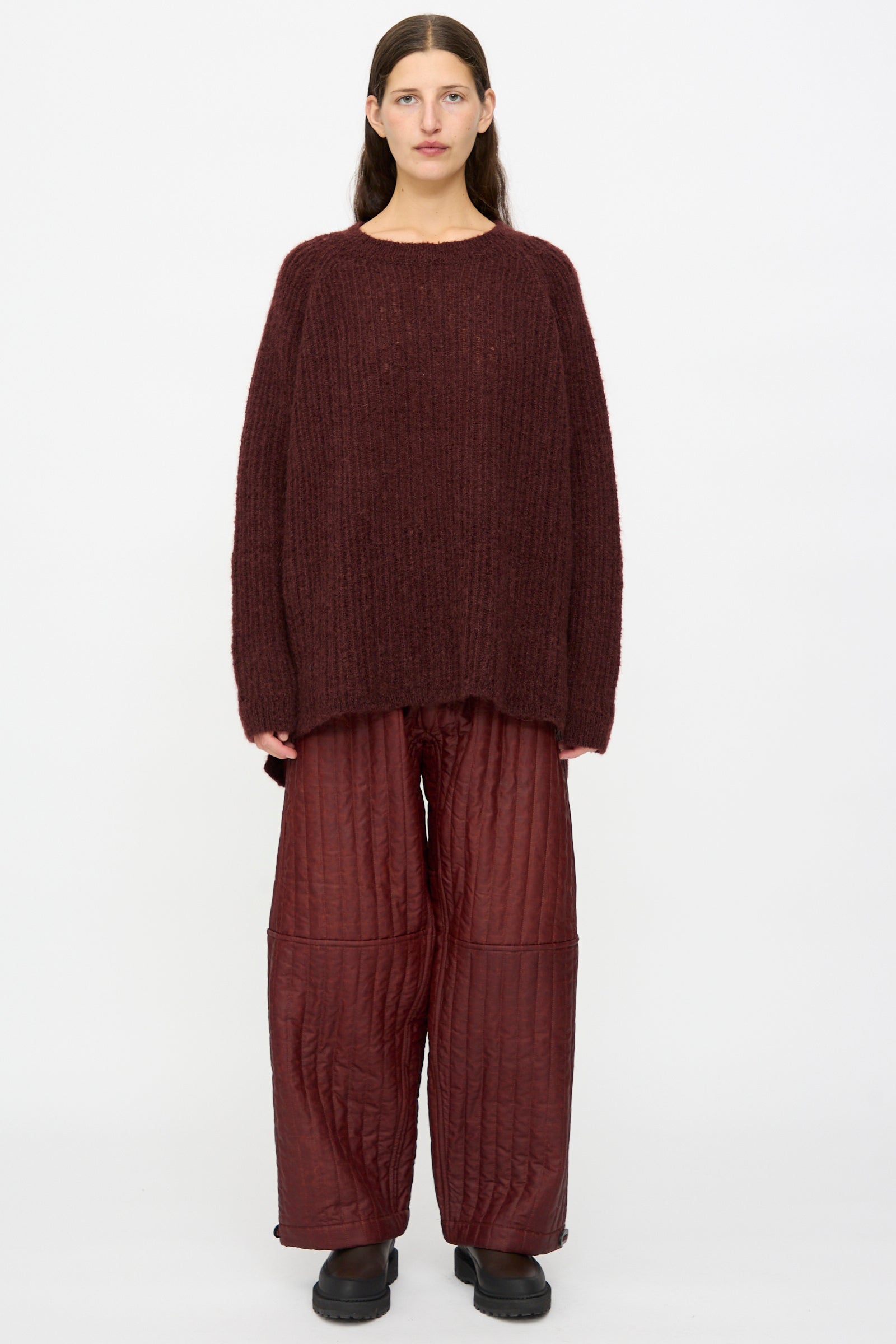 A person stands against a white background wearing the Jan-Jan Van Essche Mohair Knit #71 Sweater in Oxblood, loose quilted maroon pants, and black shoes.