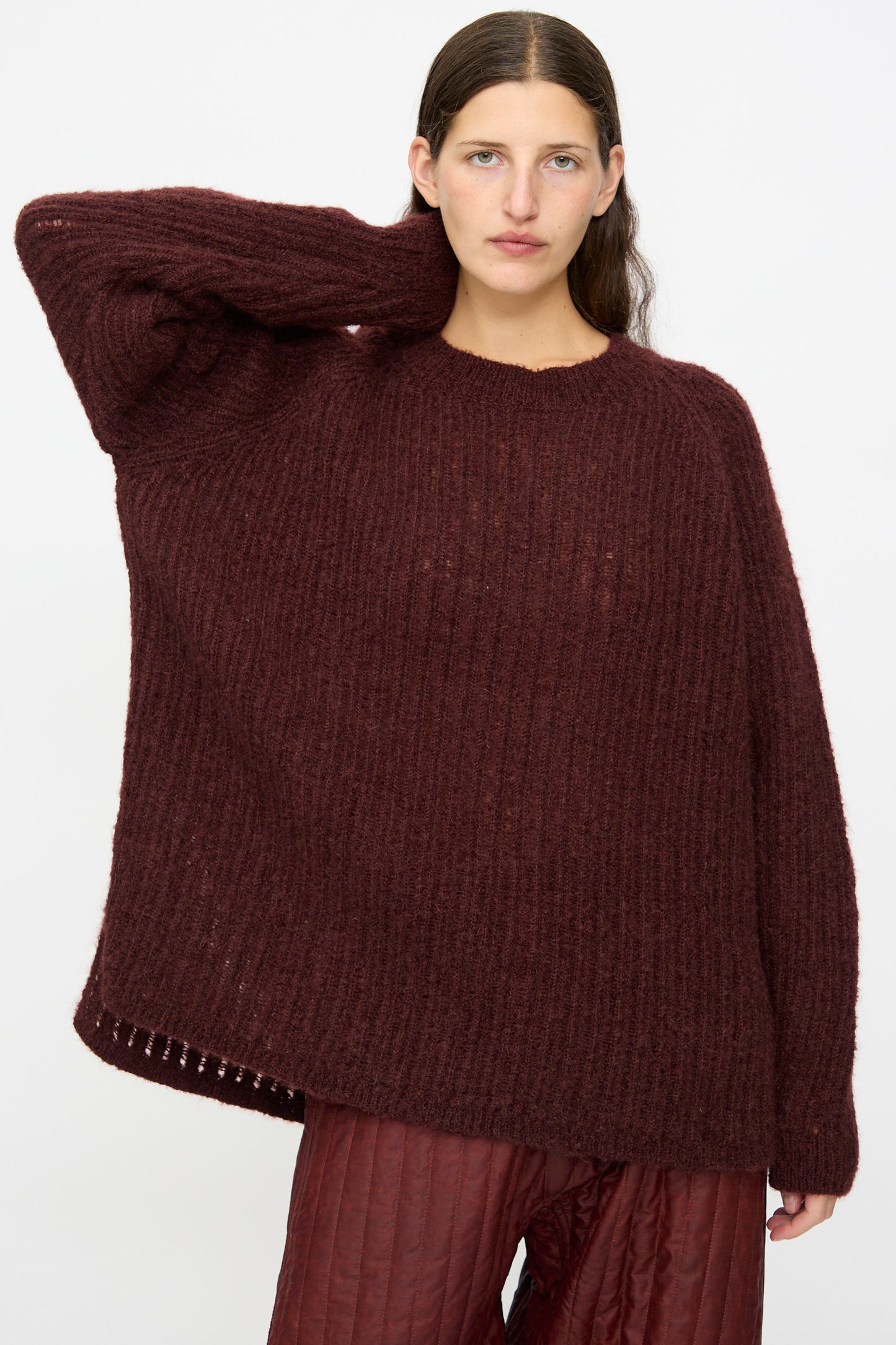 Someone stands against a plain light background wearing the Jan-Jan Van Essche Mohair Knit #71 Sweater in Oxblood paired with maroon quilted pants, one hand resting behind their head.