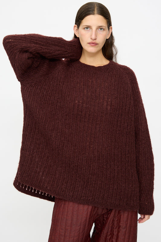 Someone stands against a plain light background wearing the Jan-Jan Van Essche Mohair Knit #71 Sweater in Oxblood paired with maroon quilted pants, one hand resting behind their head.