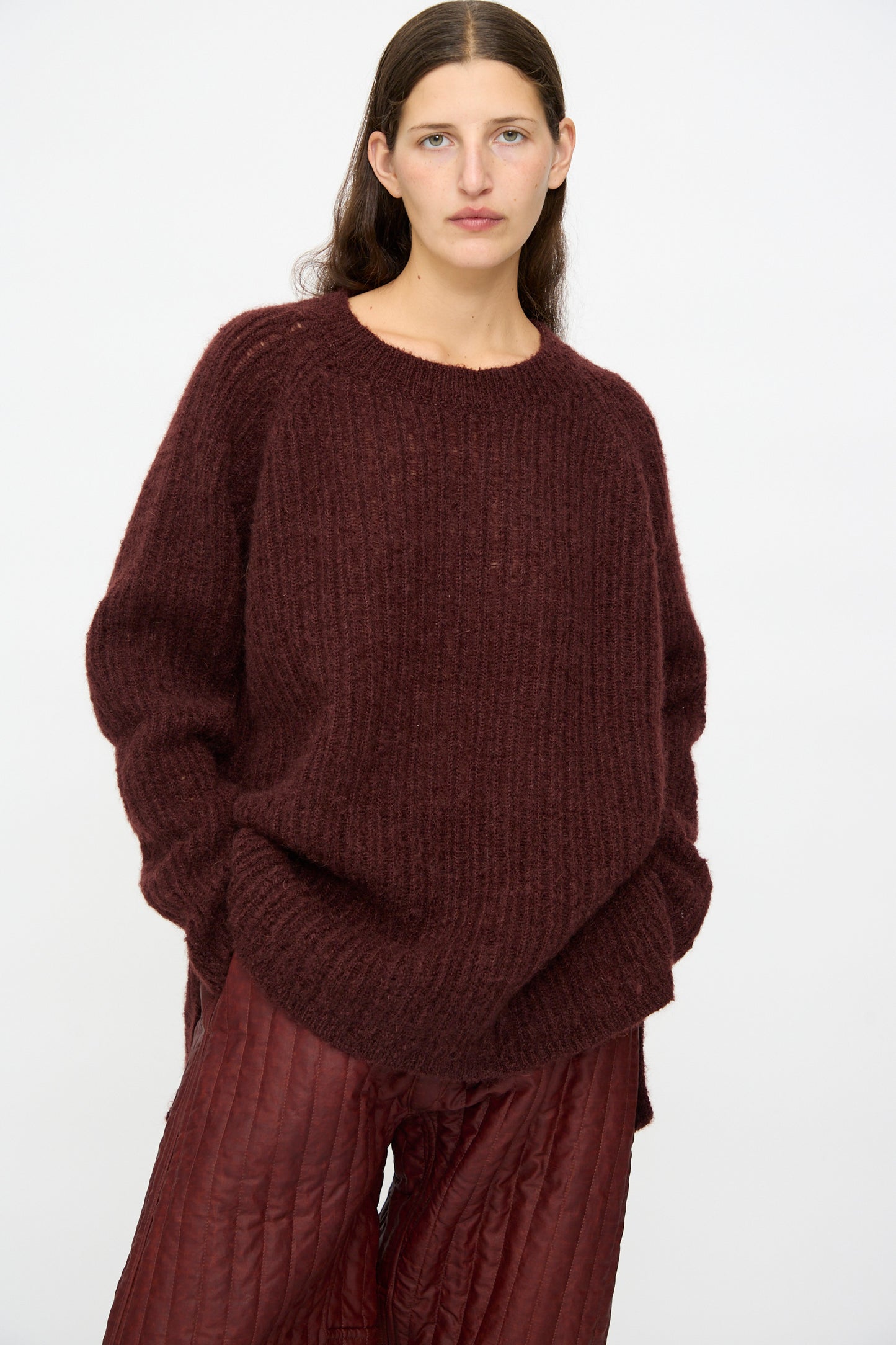 A woman with straight brown hair wears the Jan-Jan Van Essche Mohair Knit #71 Sweater in Oxblood, paired with quilted maroon pants, standing against a plain white background.
