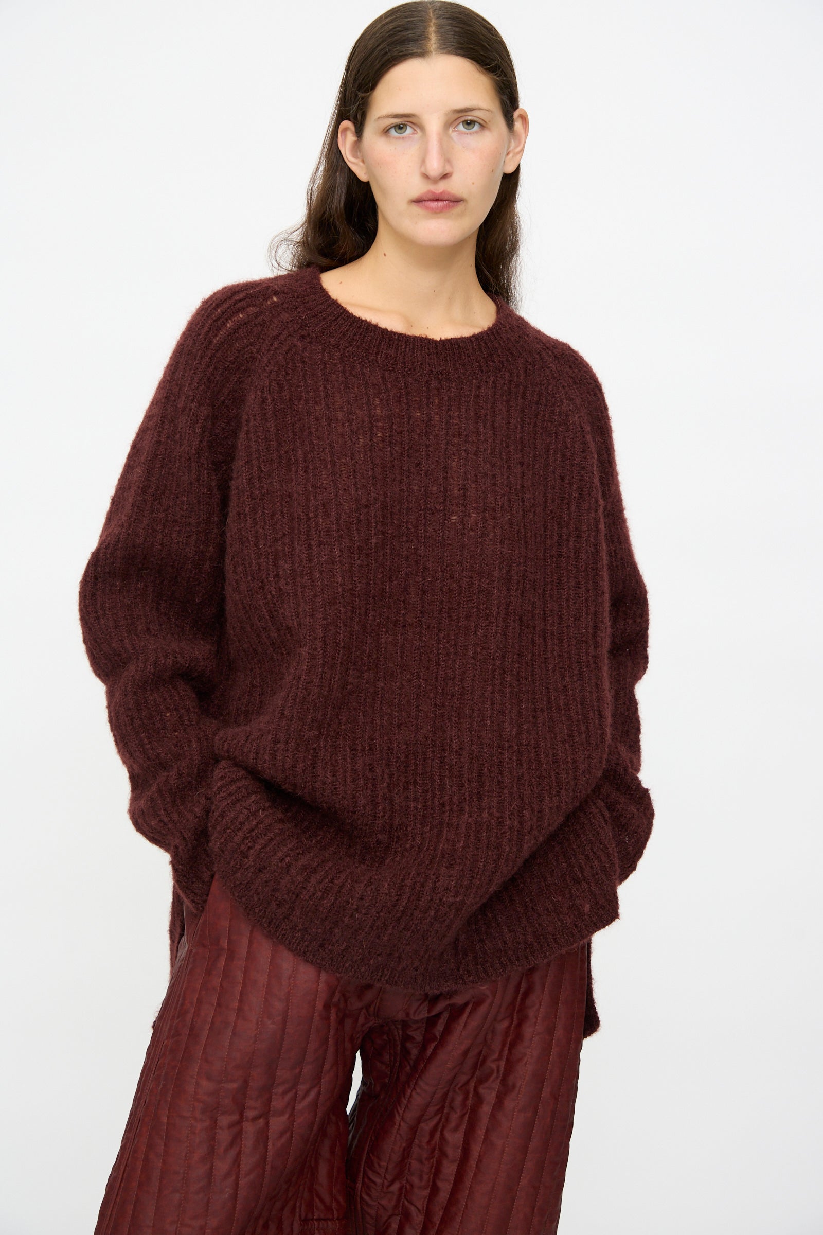 A woman with straight brown hair wears the Jan-Jan Van Essche Mohair Knit #71 Sweater in Oxblood, paired with quilted maroon pants, standing against a plain white background.