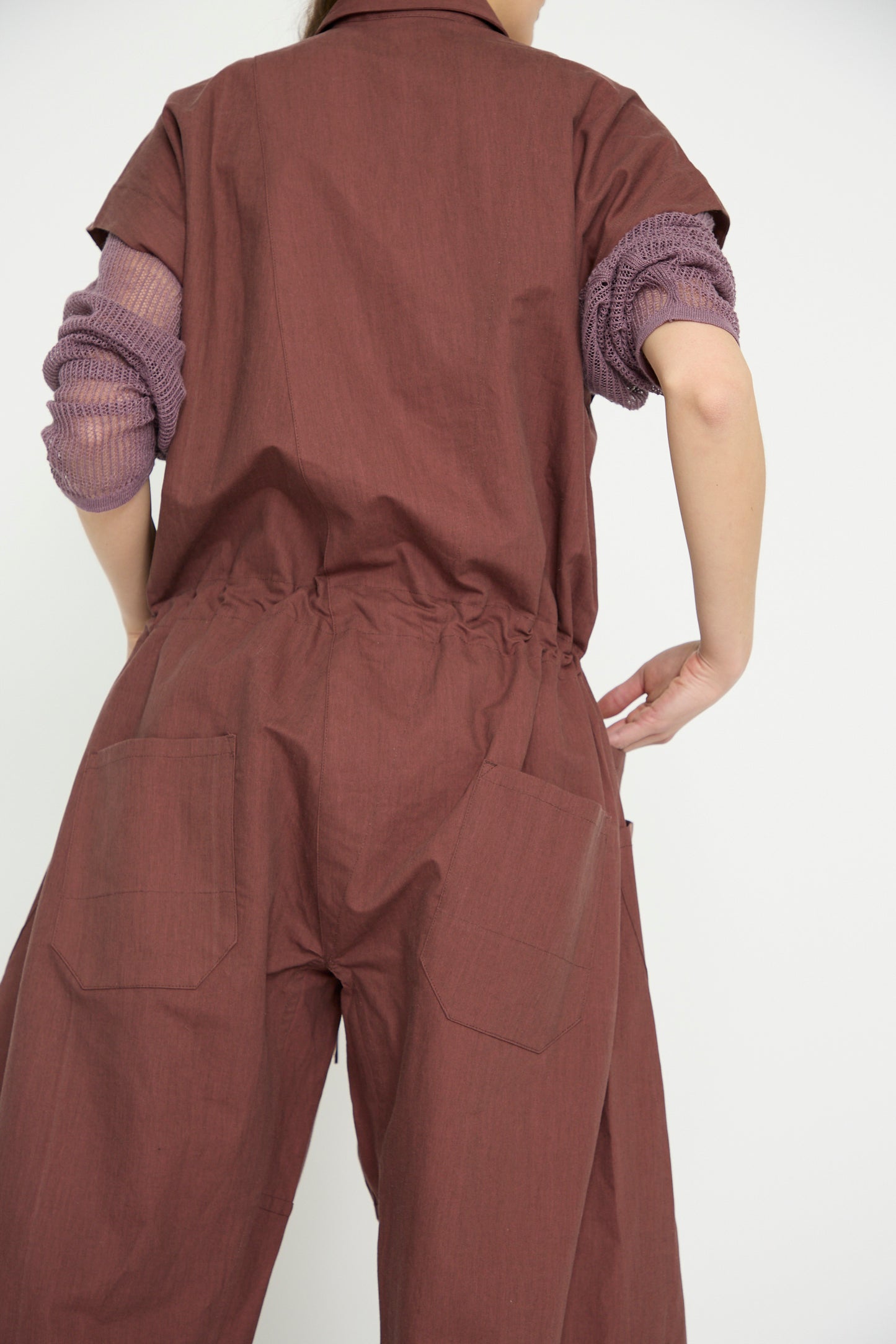 The individual, seen from behind, wears the Oxford Cloth Jumpsuit #12 in Prune by Jan-Jan Van Essche, complemented by a purple long-sleeve top. The pristine white background accentuates the ensemble's casual elegance.