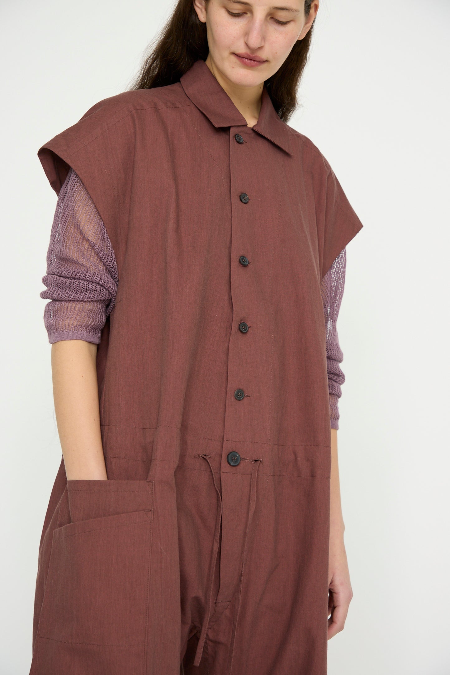 A person stands wearing Jan-Jan Van Essche's sleeveless brown Oxford Cloth Jumpsuit #12 in Prune over a purple long-sleeve top with a pointed collar, hands in pockets, looking downward.