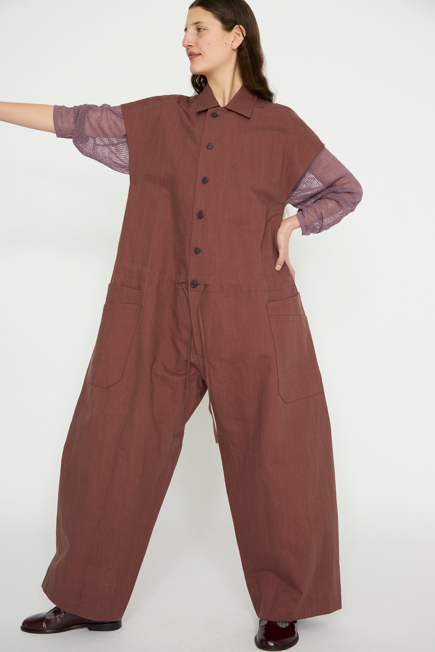 A person in an oversized Jan-Jan Van Essche Oxford Cloth Jumpsuit #12 in Prune, featuring a pointed collar and layered over a purple shirt, stands against a plain background with one arm extended to the side.