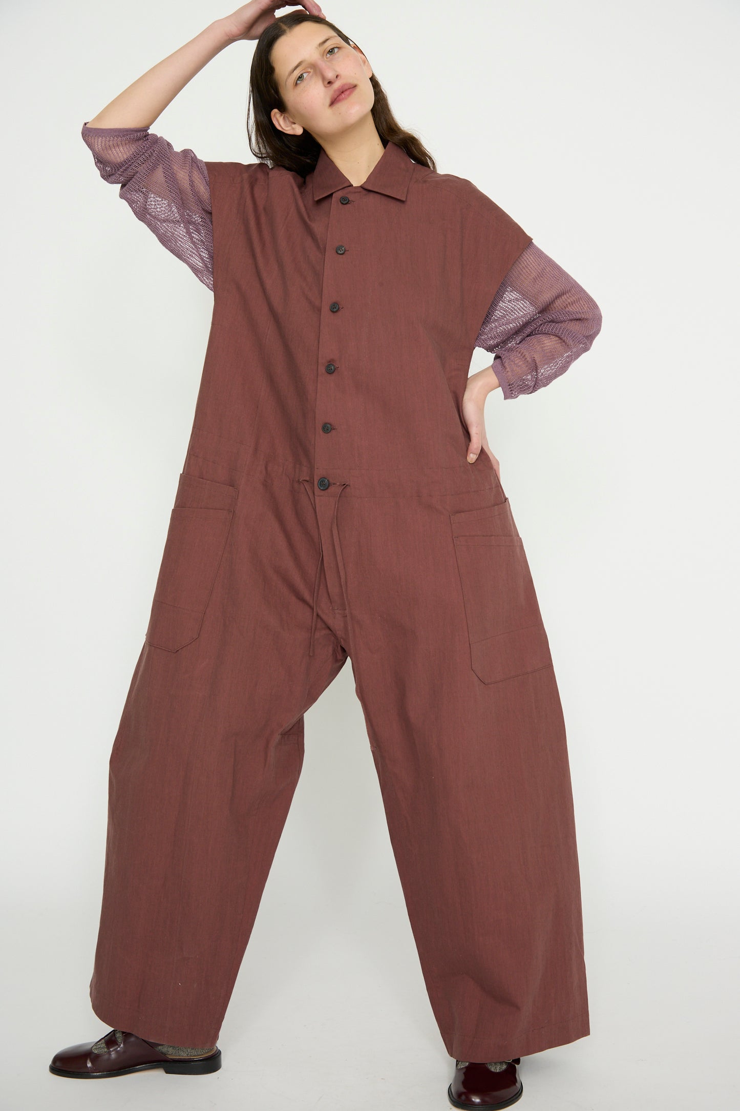 A person poses against a plain backdrop wearing Jan-Jan Van Essche's Oxford Cloth Jumpsuit #12 in Prune, crafted from a comfortable cotton blend, with a purple shirt underneath and one hand resting on their head.