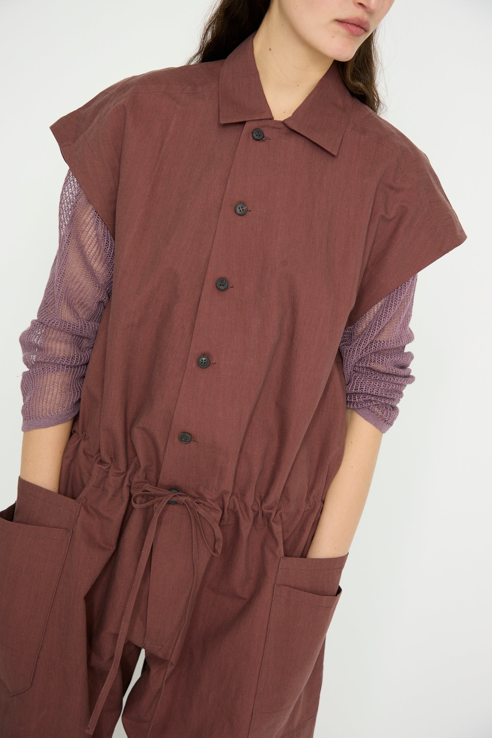 Someone is wearing the Jan-Jan Van Essche Oxford Cloth Jumpsuit #12 in Prune layered over a light purple mesh long sleeve shirt, standing against a plain background.