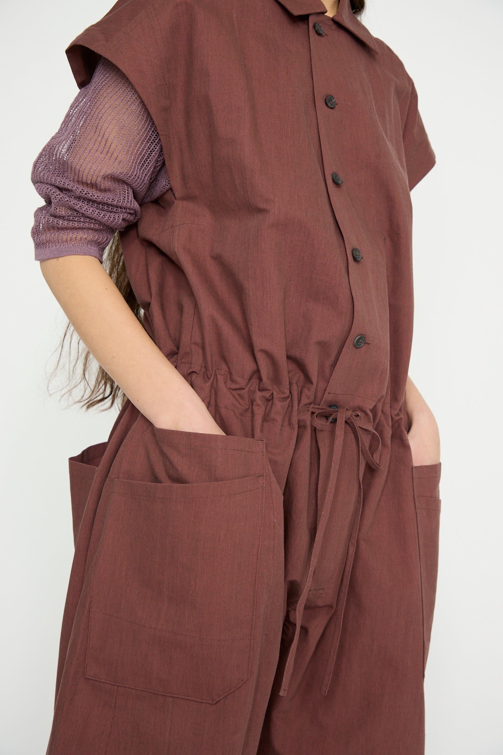 A person is seen wearing the Jan-Jan Van Essche Oxford Cloth Jumpsuit #12 in Prune, featuring a button-up front, pointed collar, and drawstring waist over a sheer long-sleeve top. The jumpsuit's design includes large pockets for a practical yet stylish look.