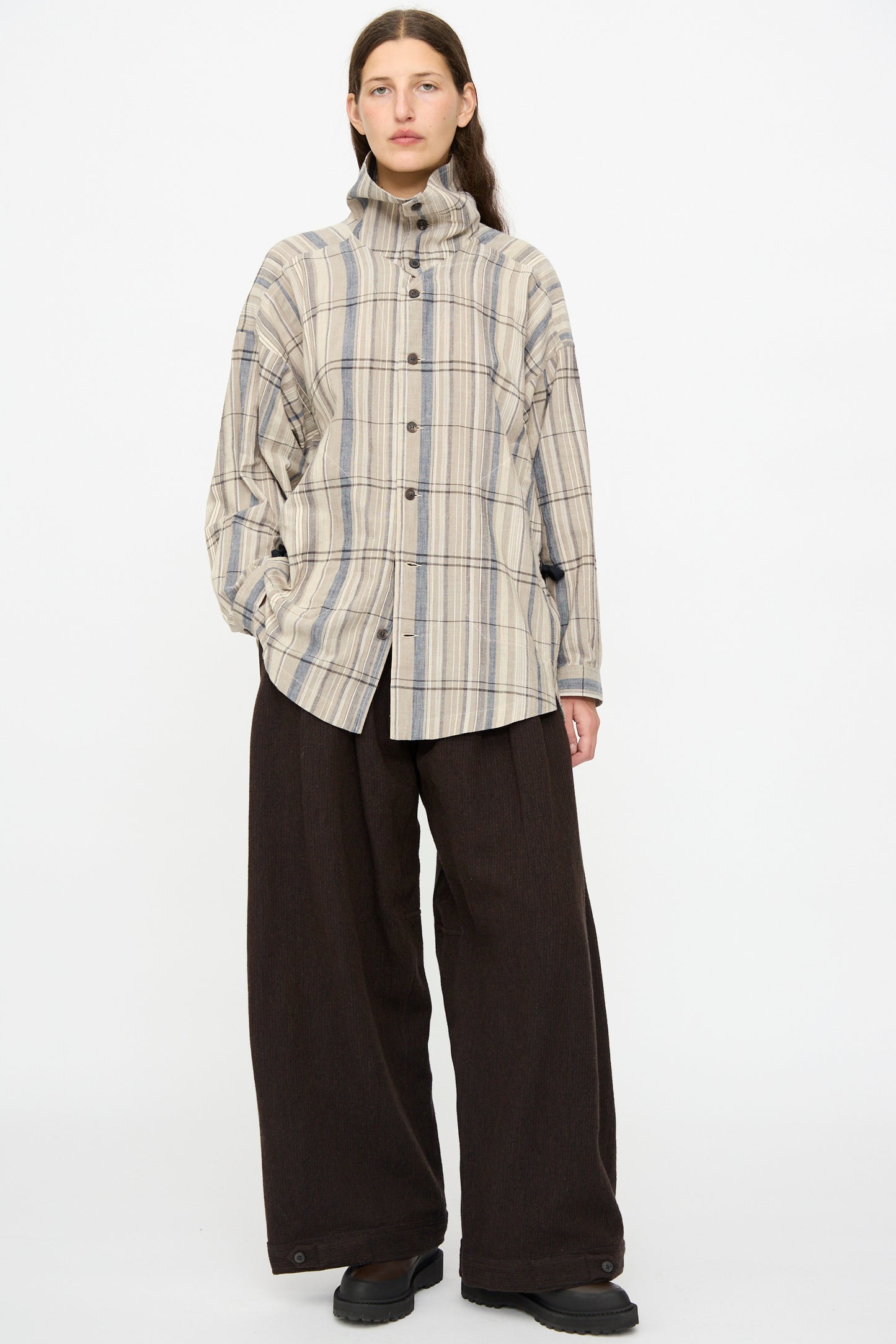 A person faces forward wearing Jan-Jan Van Essche Ridged Linen Wool Trousers #87 in Indigo and Eggplant, paired with a beige plaid button-up shirt with a high collar.