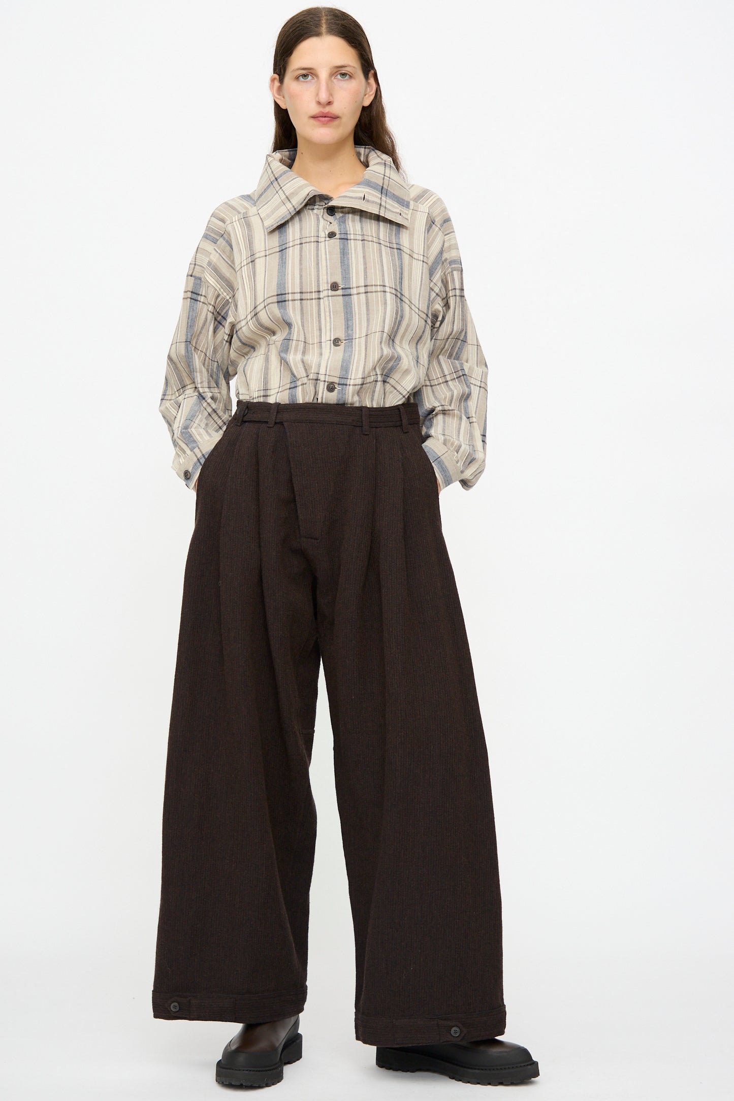 Person standing against a white background, wearing a beige plaid button-up shirt tucked into Jan-Jan Van Essche Ridged Linen Wool Trousers #87 in Eggplant and indigo, paired with black shoes.
