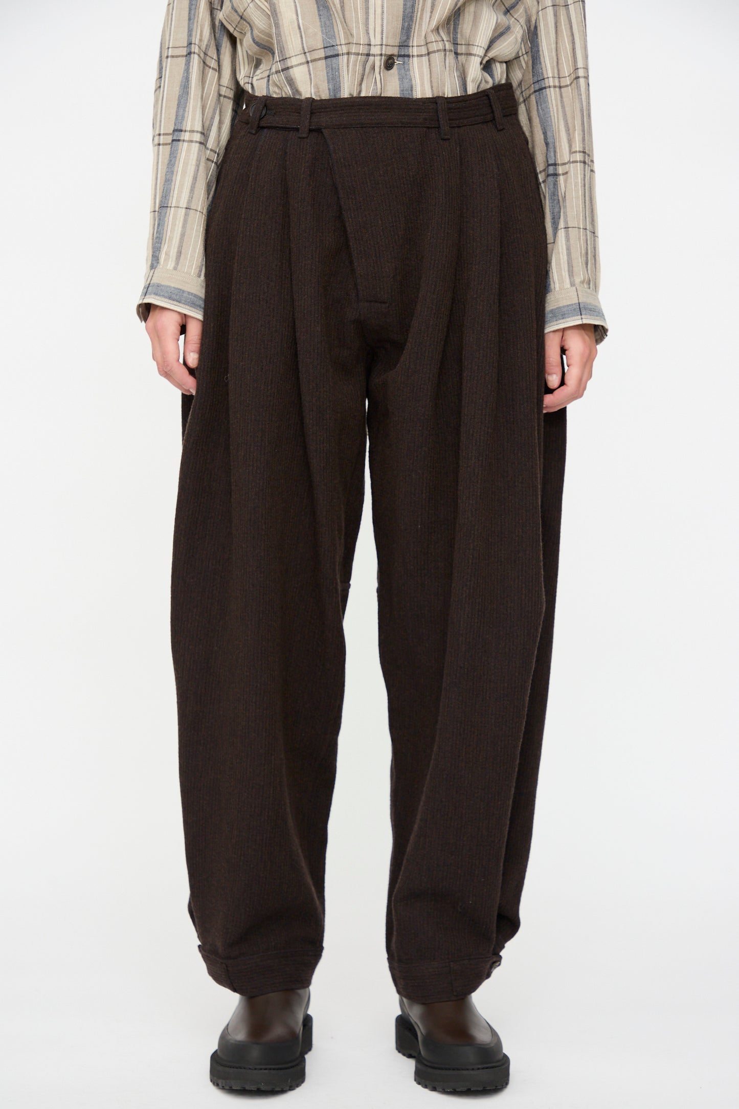 A person wears Jan-Jan Van Essche Ridged Linen Wool Trousers #87 in Eggplant, paired with a beige plaid shirt and dark shoes, standing against a plain white background.