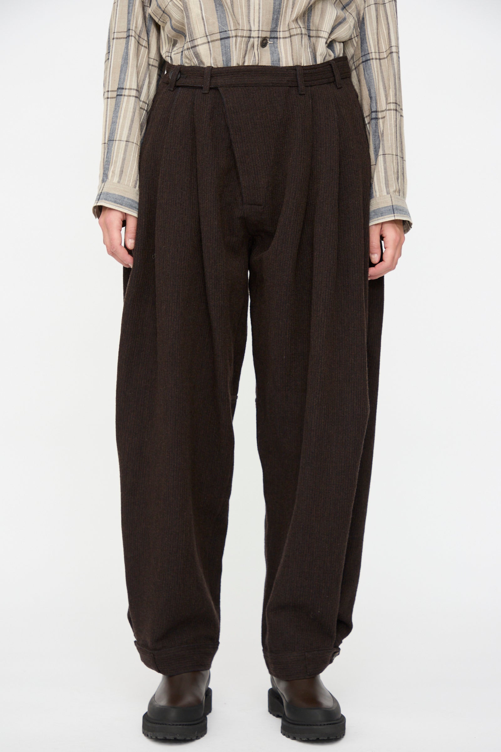 A person wears Jan-Jan Van Essche Ridged Linen Wool Trousers #87 in Eggplant, paired with a beige plaid shirt and dark shoes, standing against a plain white background.