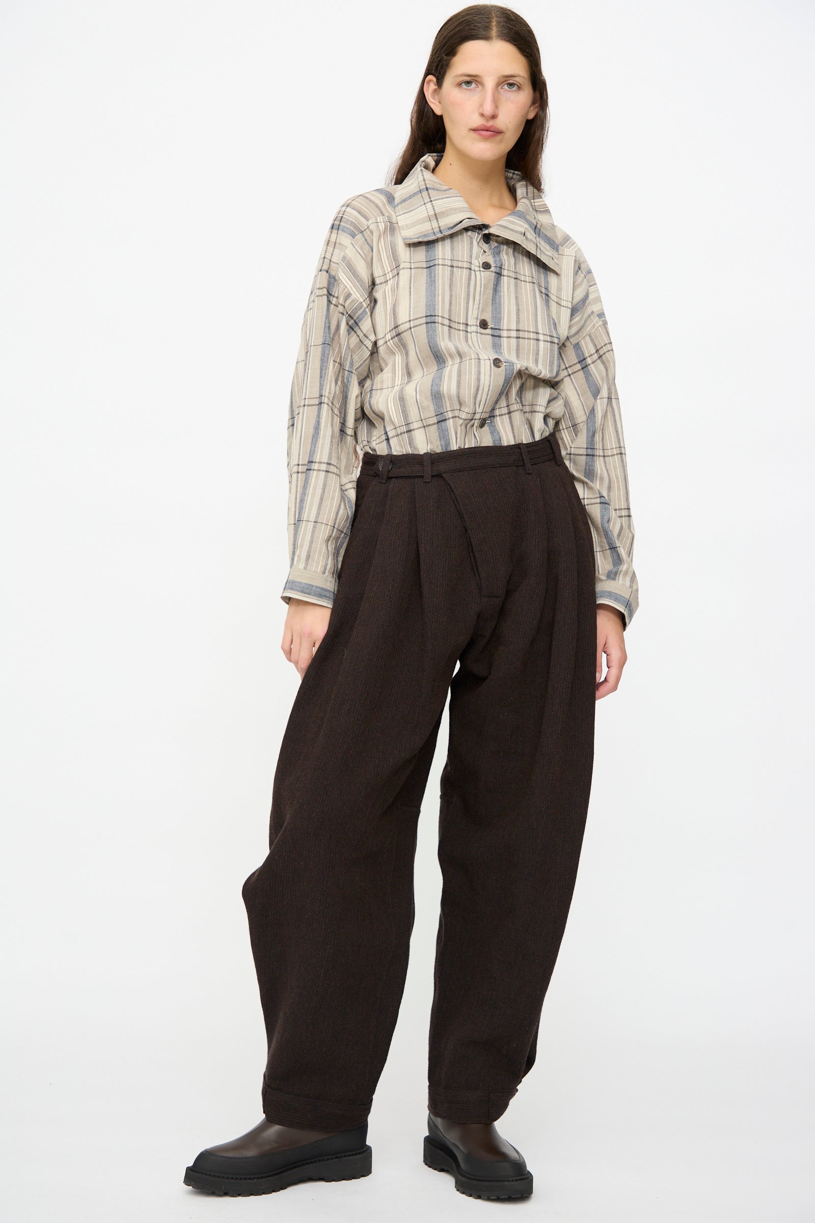A person stands against a plain white background wearing a beige and blue plaid button-up shirt, Jan-Jan Van Essche Ridged Linen Wool Trousers #87 in Indigo and Eggplant, and black boots.