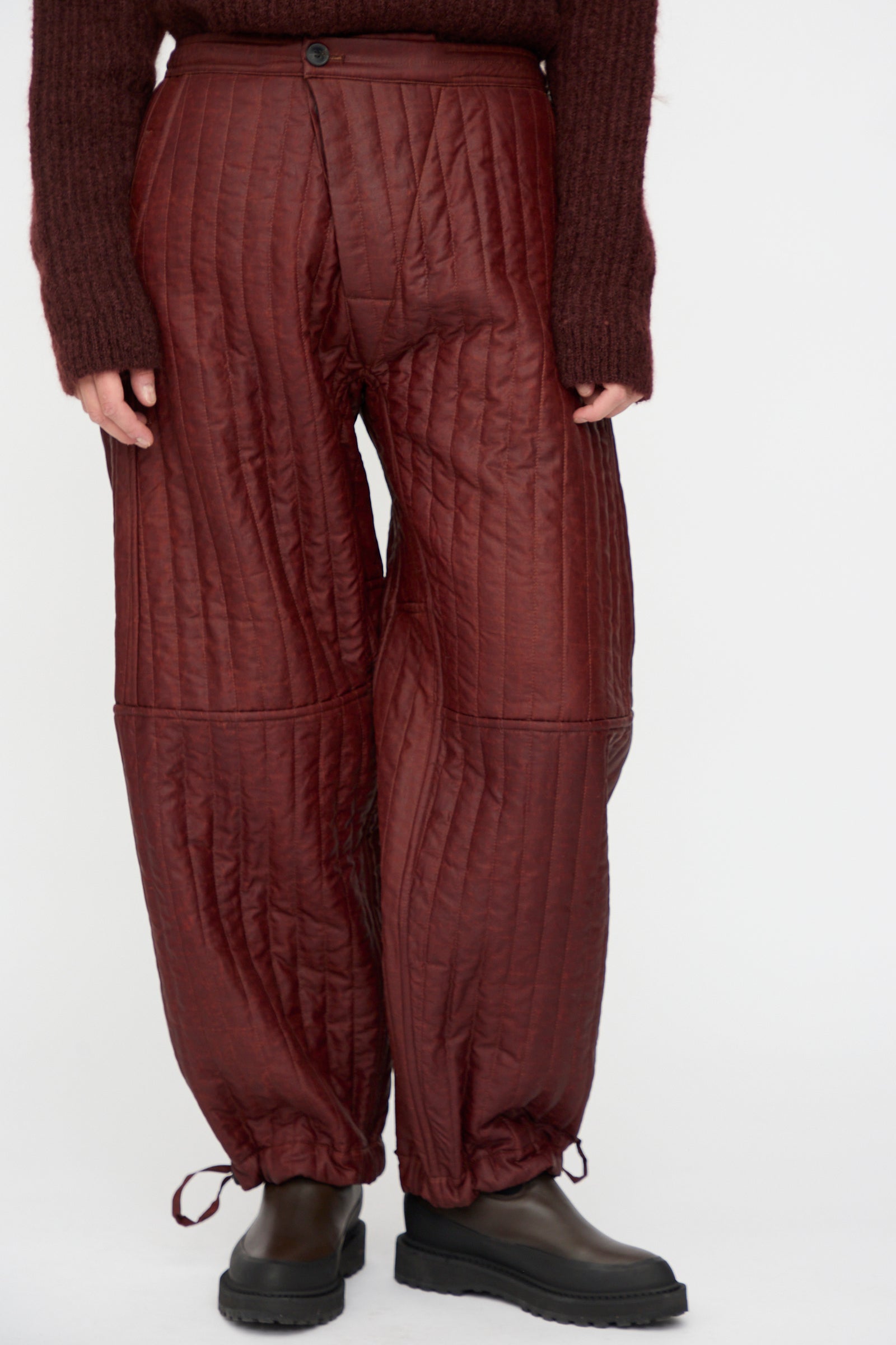 A person wears Jan-Jan Van Essche's Shuliang Silk Padded Trousers #89 in oxblood, styled with a brown sweater and black shoes; image is cropped at the shoulders.