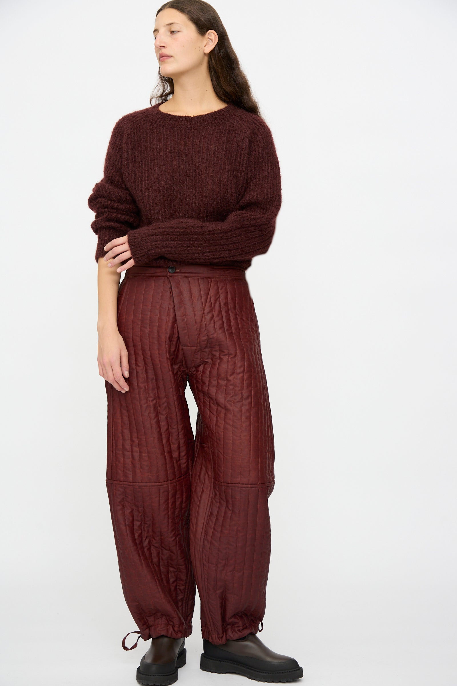 A woman stands against a plain background wearing a textured burgundy sweater, Jan-Jan Van Essche Shuliang Silk Padded Trousers #89 in Oxblood, and black boots.