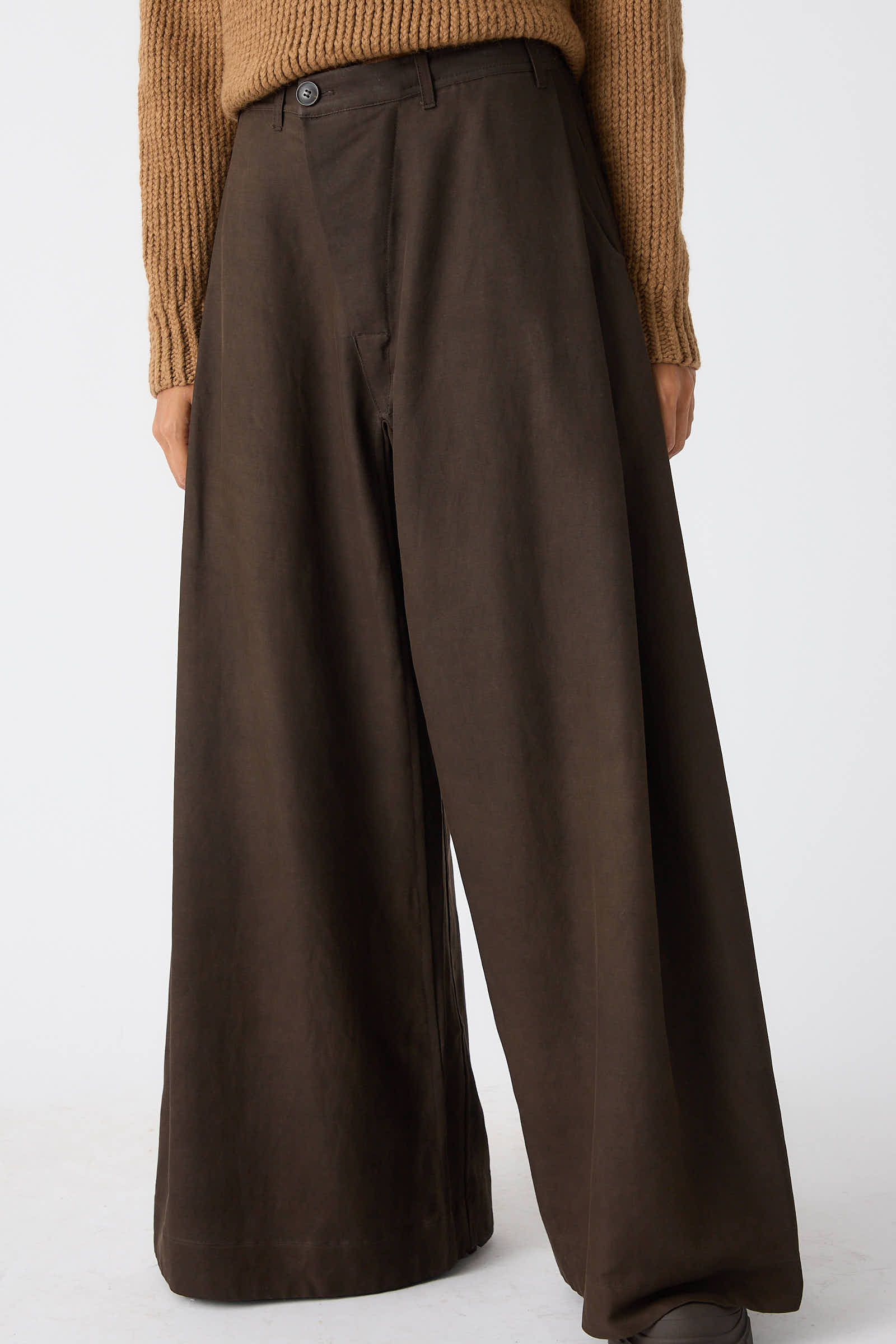 A person wearing a brown knit sweater and Jan-Jan Van Essche's Silk Wool Twill Trouser in Amami Mud stands against a plain white background. Only the lower half of their body, showcasing the side tuck detail on the relaxed fit, wide-leg trousers is visible.