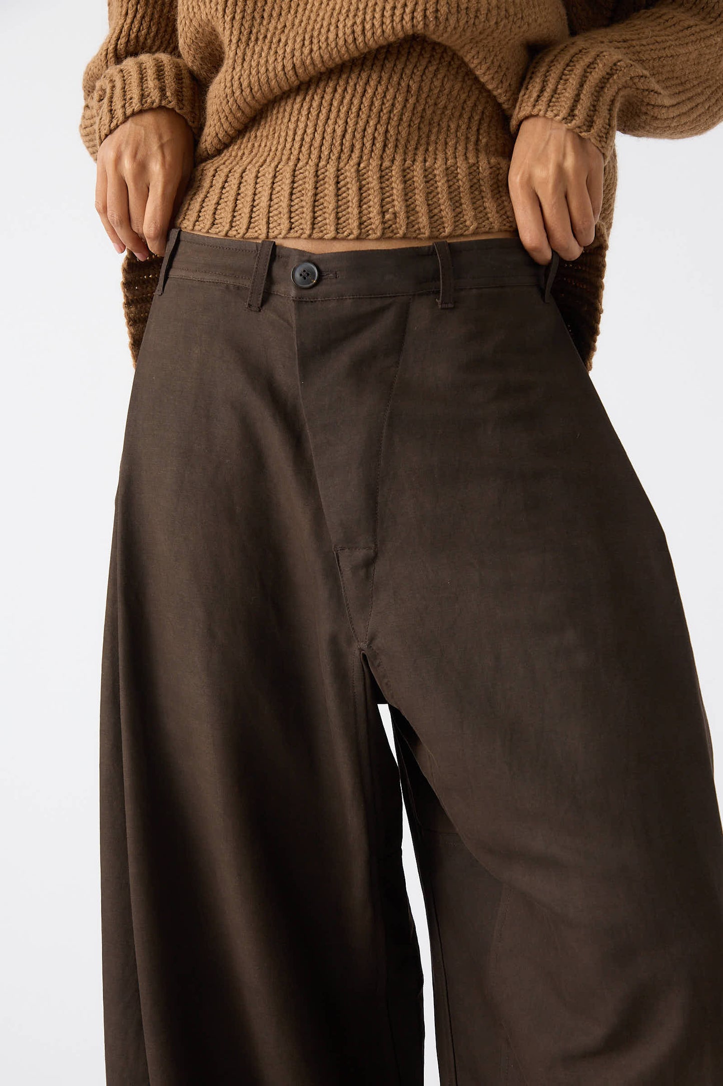 A person in a tan knitted sweater is shown adjusting or pulling up a pair of Jan-Jan Van Essche Silk Wool Twill Trouser in Amami Mud, which feature a relaxed fit and side tuck detail.