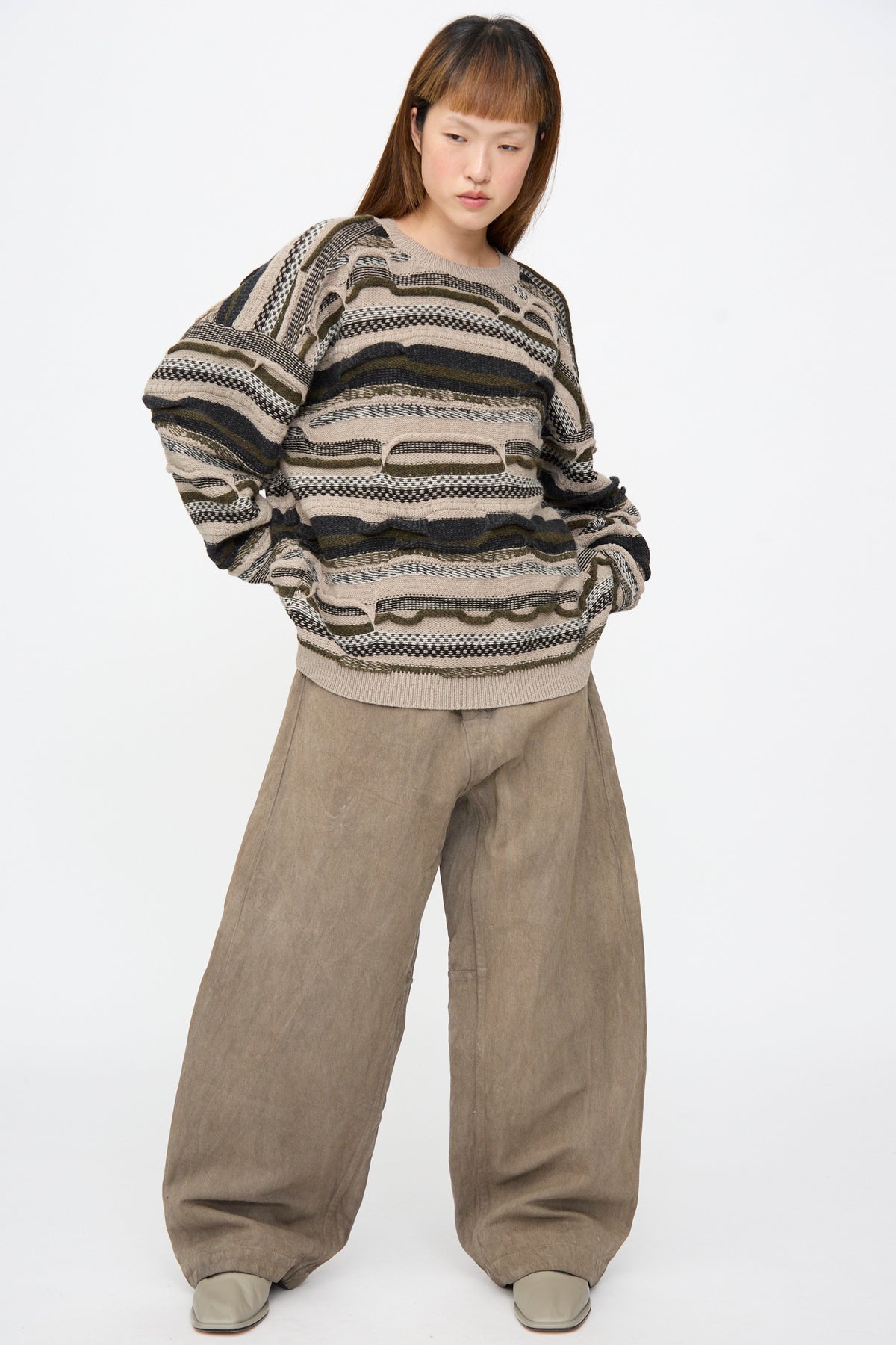 A person wears an oversized, multi-textured Super Fine Wool Knit Sweater #80 in Light Variation with horizontal stripes in tan, olive, and black, paired with wide-leg beige trousers and grey shoes.