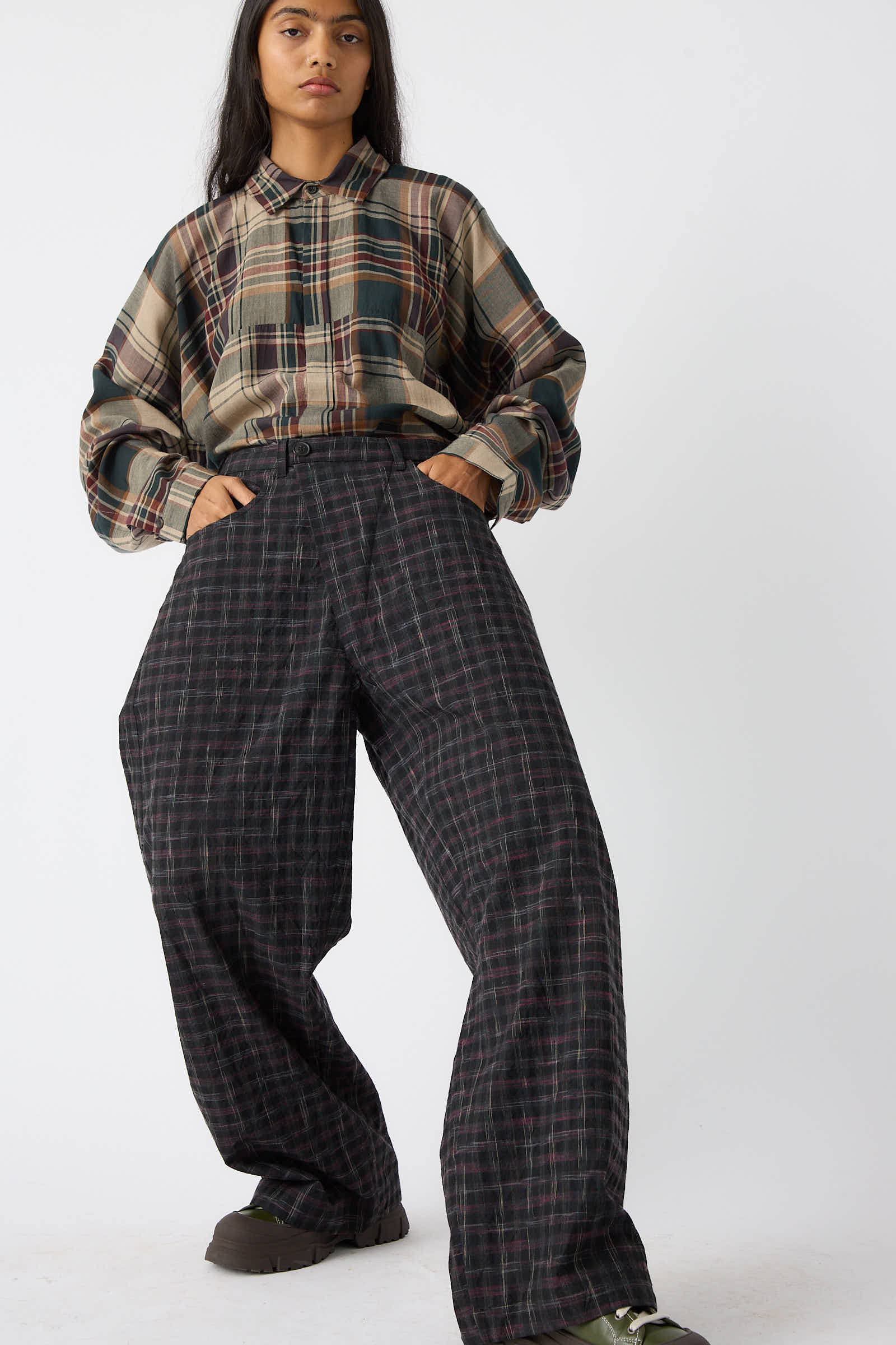 A person dressed in a plaid shirt and the Tumbled Wool Check Trouser in Kasuri by Jan-Jan Van Essche, with a relaxed fit and oversized style, stands against a plain white background with hands in pockets.