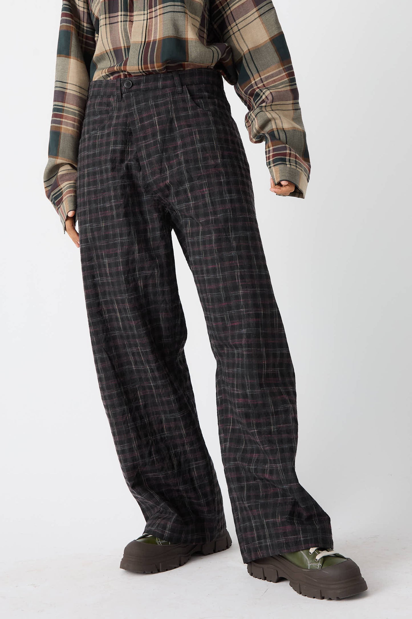 A person wearing a plaid shirt, paired with the Tumbled Wool Check Trouser in Kasuri from Jan-Jan Van Essche, stands against a plain background. Green-and-black sneakers complete the look.