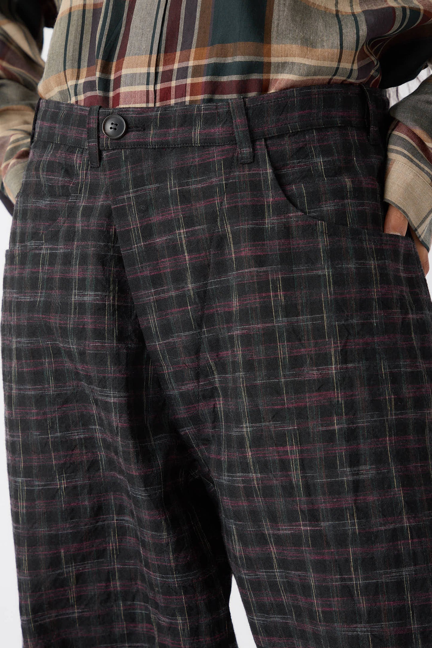 Close-up of a person's midsection wearing a multicolored plaid shirt and Jan-Jan Van Essche's Tumbled Wool Check Trouser in Kasuri with hands in pockets, showcasing a relaxed fit.