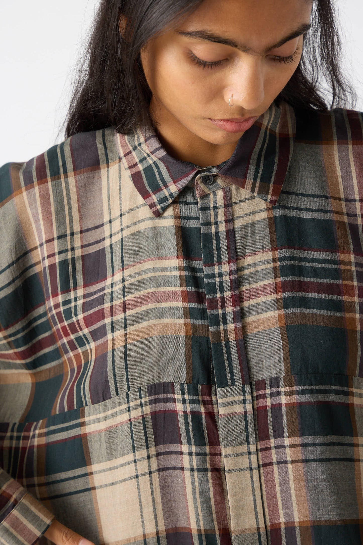 A person with long dark hair looks down while wearing a loose fit Jan-Jan Van Essche Viscose Shirt in Sand, Green, and Burgundy madras plaid patterns.