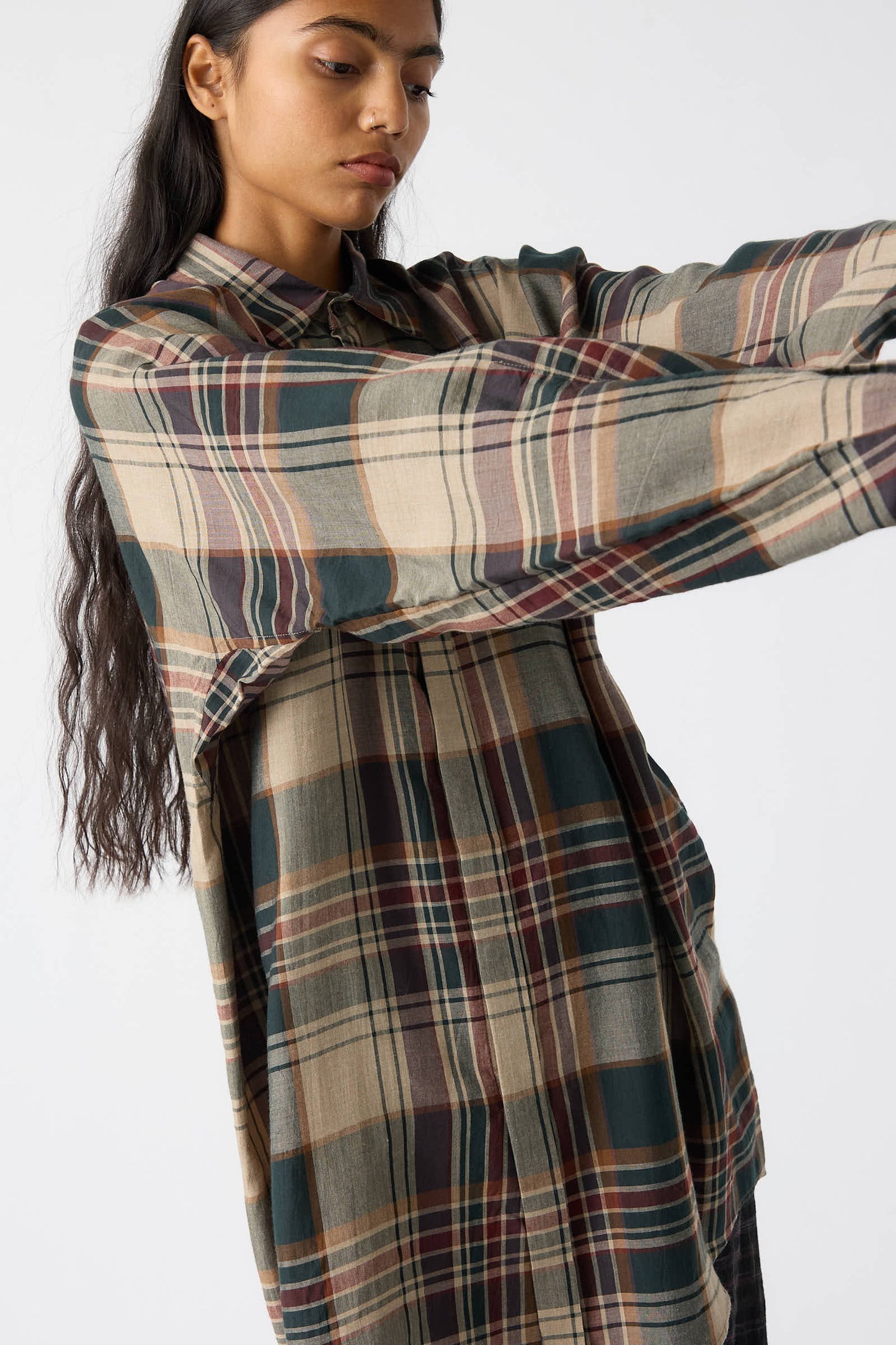 A person with long dark hair wears an oversized Jan-Jan Van Essche viscose shirt in sand, green, and burgundy, and stretches their arms forward against a plain background.