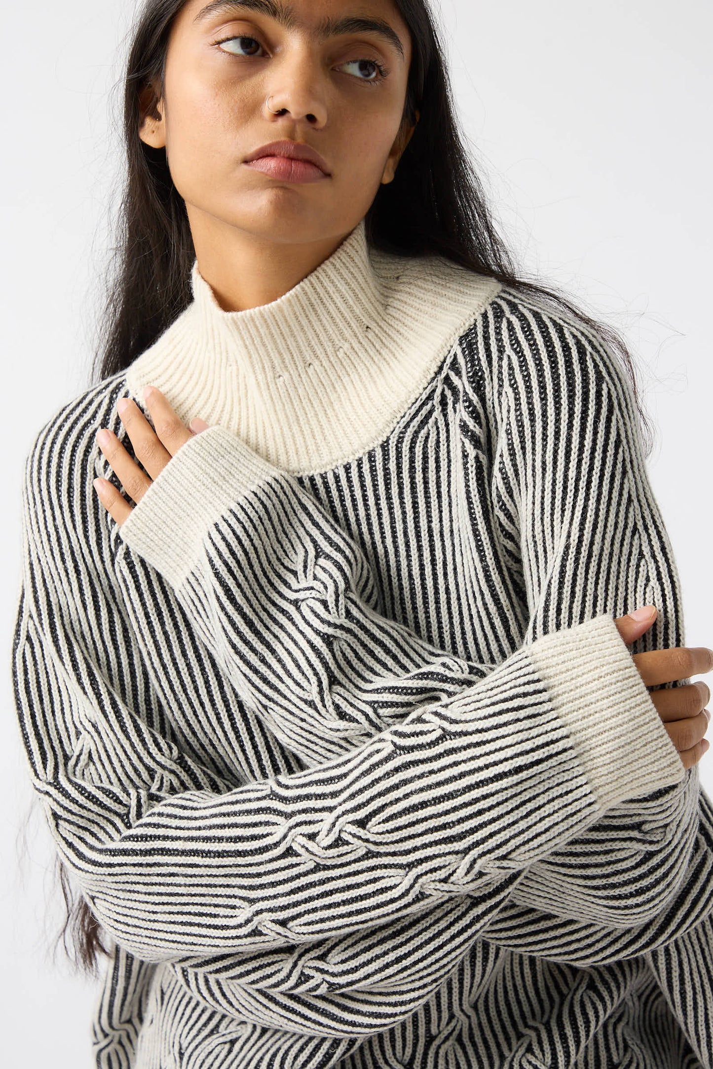 A person with long hair wears a black-and-white striped loose fit sweater and the Jan-Jan Van Essche Wool Linen Cable Knit Sweater in White and Anthracite, crossing their arms with a neutral expression.