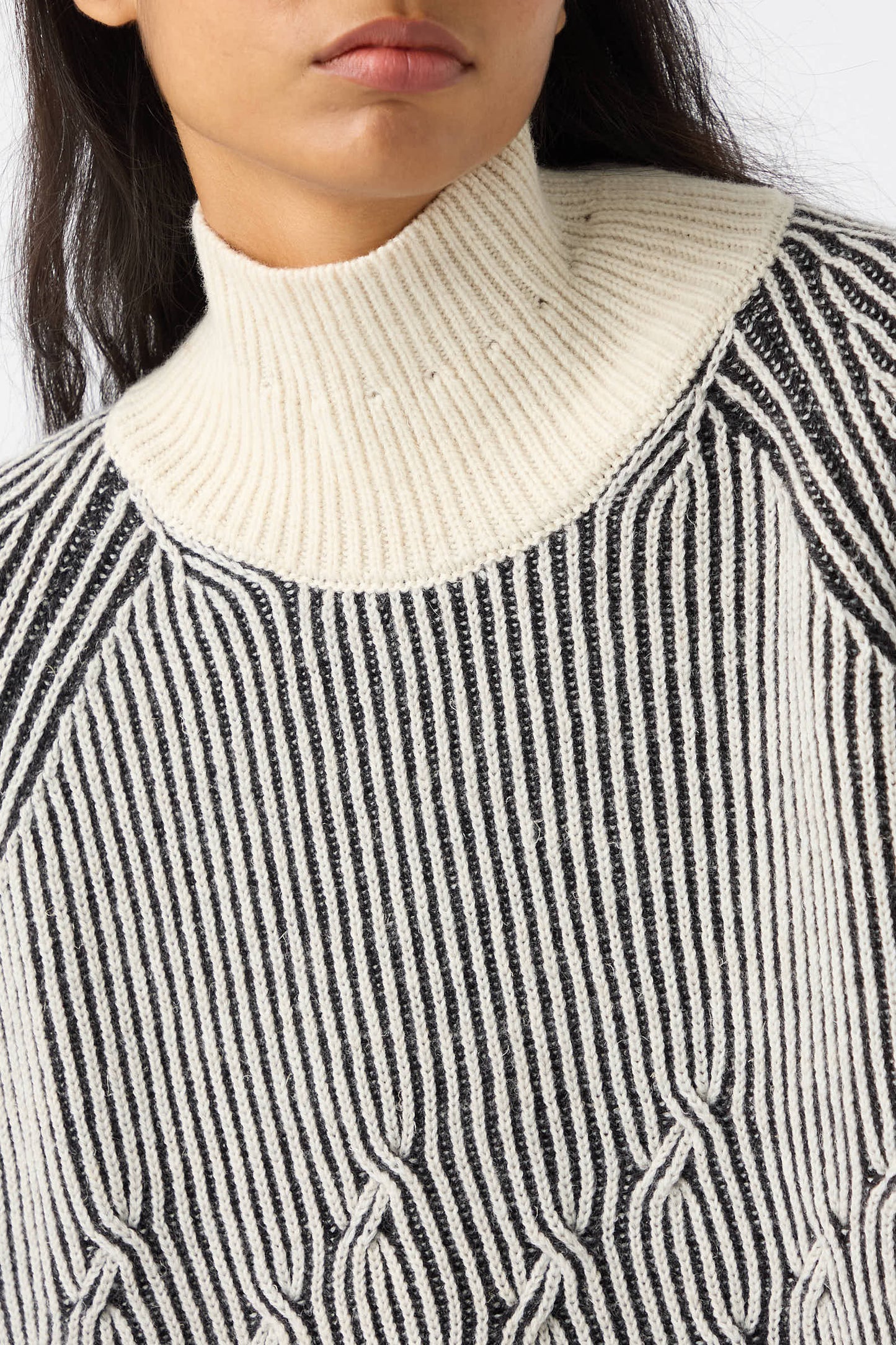 A close-up of a person wearing the Jan-Jan Van Essche Wool Linen Cable Knit Sweater in White and Anthracite. The high-neck, black and white striped sweater features intricate cable knit patterns and a loose fit, made from a cozy wool/linen blend that perfectly complements the person's long, dark hair.