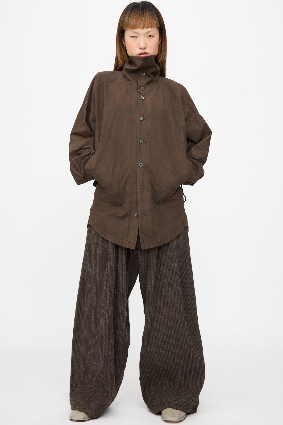 A person wearing a brown button-up jacket and Jan-Jan Van Essche Yak Linen Cotton Trousers #93 in Brown Striped, which feature an extra-wide, pinstriped design with a double-pleated front.