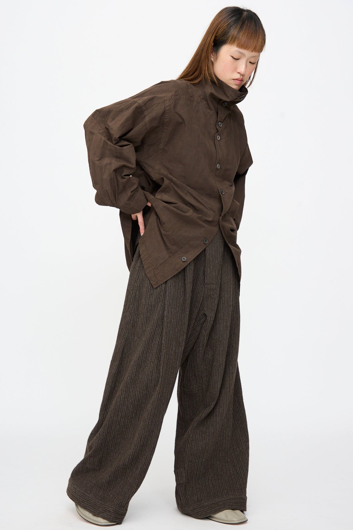 A person wearing a brown button-up shirt and Jan-Jan Van Essche Yak Linen Cotton Trousers #93 in Brown Striped, featuring an extra-wide, double-pleated design.