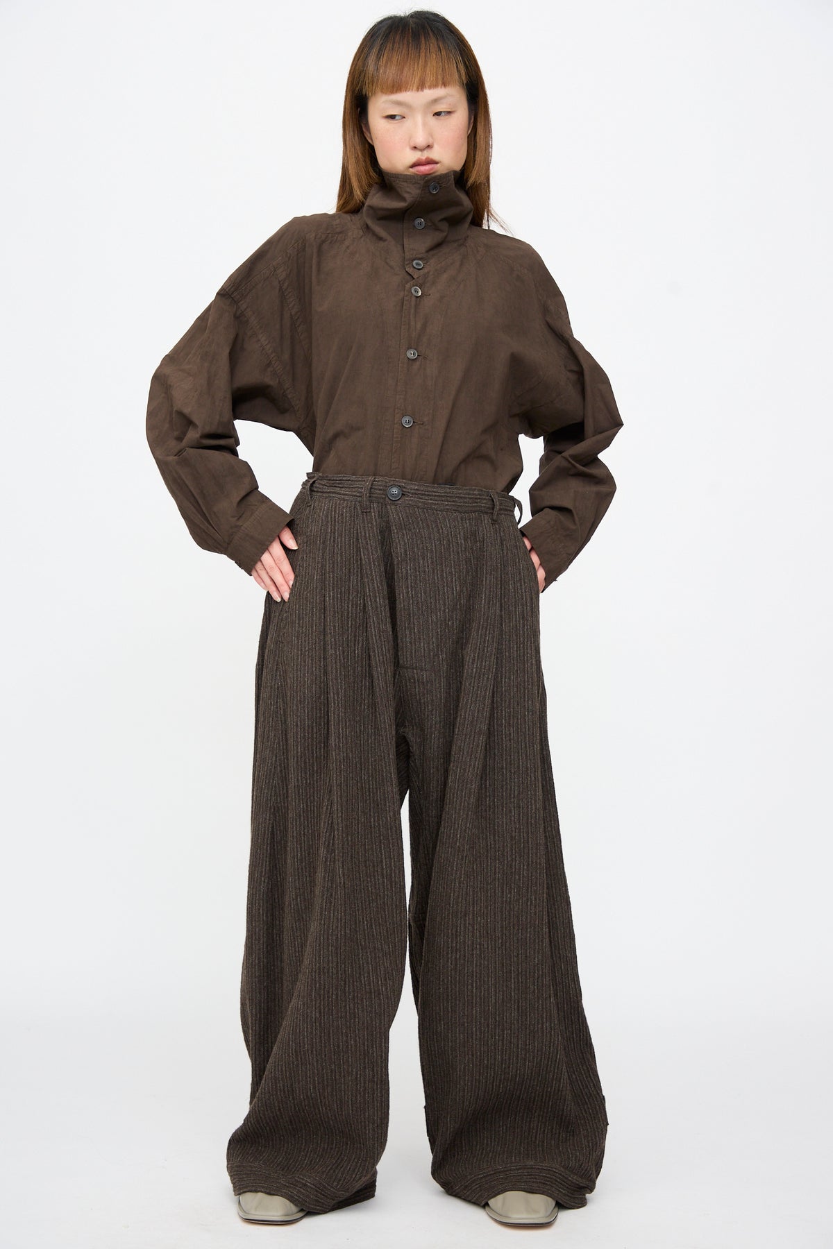 A person wearing a brown high-neck shirt and Jan-Jan Van Essche Yak Linen Cotton Trousers #93 in Brown Striped, featuring a double-pleated, extra-wide silhouette with subtle vertical stripes.