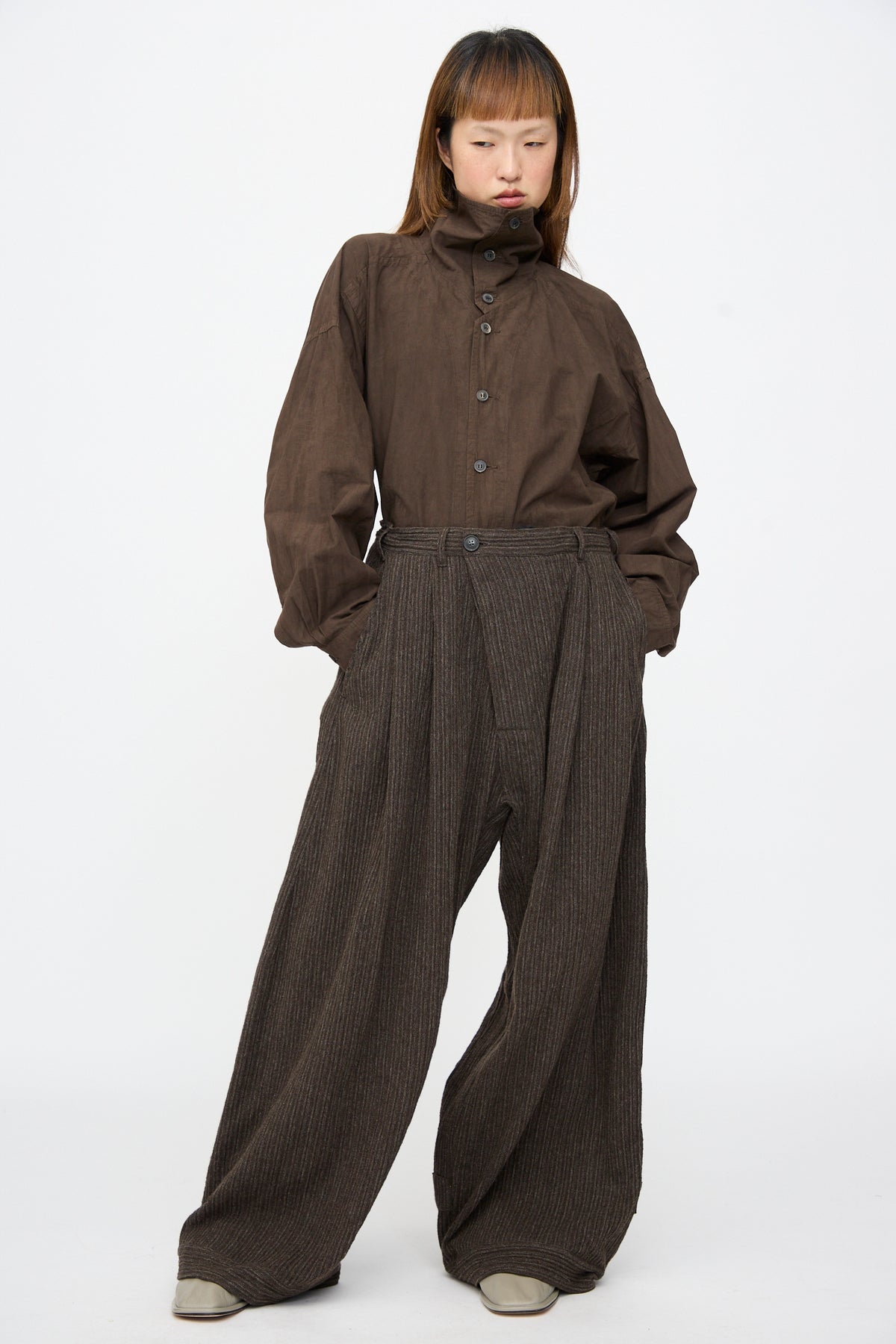 A person wearing a dark brown button-up shirt and Jan-Jan Van Essche Yak Linen Cotton Trousers #93 in Brown Striped, which are extra wide and double-pleated, with hands in pockets.